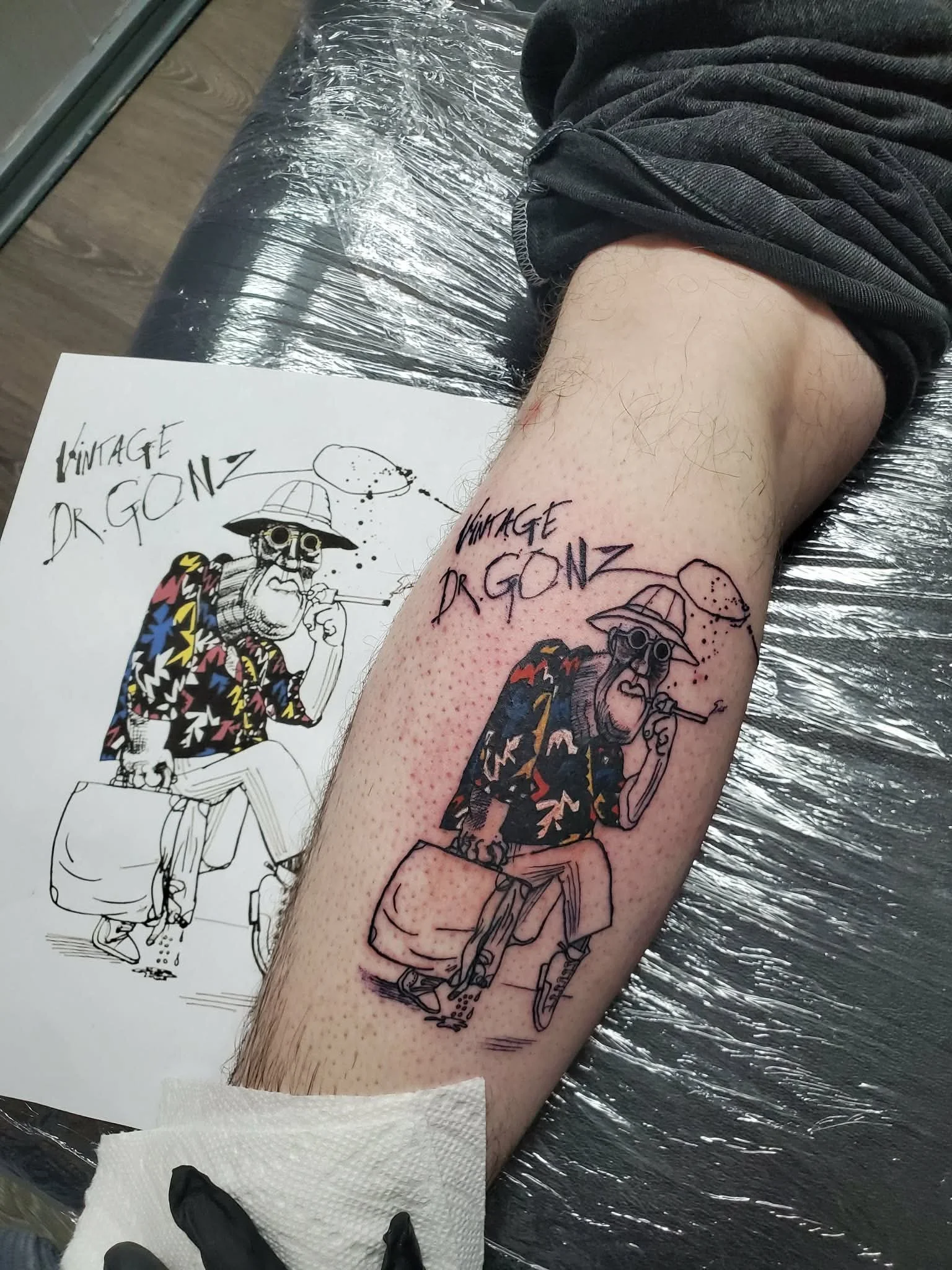 A person is getting a tattoo of a cartoon character sitting on a bench, smoking a pipe, wearing a hat, sunglasses, a colorful Hawaiian shirt, and striped pants. The tattoo says 'VINTAGE DR. GONZ' above the character.