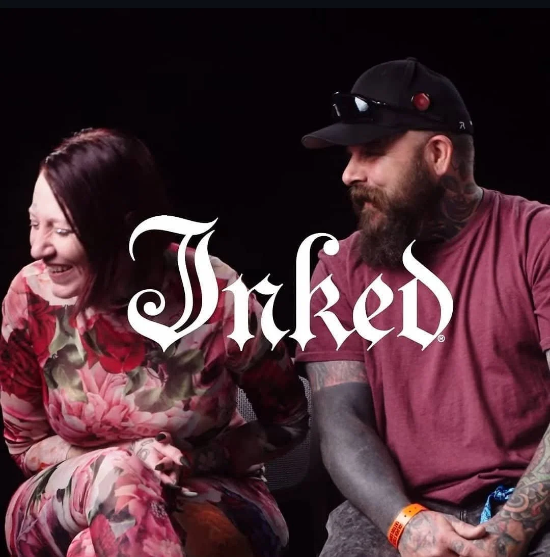 A woman with dark red hair laughing, wearing a floral dress, sitting next to a tattooed man in a maroon t-shirt and black cap with sunglasses. The word 'Taked' is overlaid on the image in a gothic font.