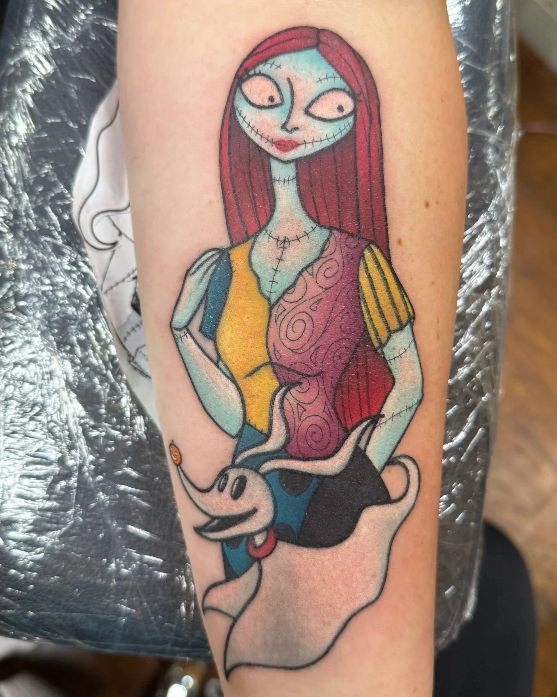 Tattoo of Nightmare Before Christmas character Sally, stitched skin, and a colorful dress, accompanied by a cartoon dog Zero with a collar.
