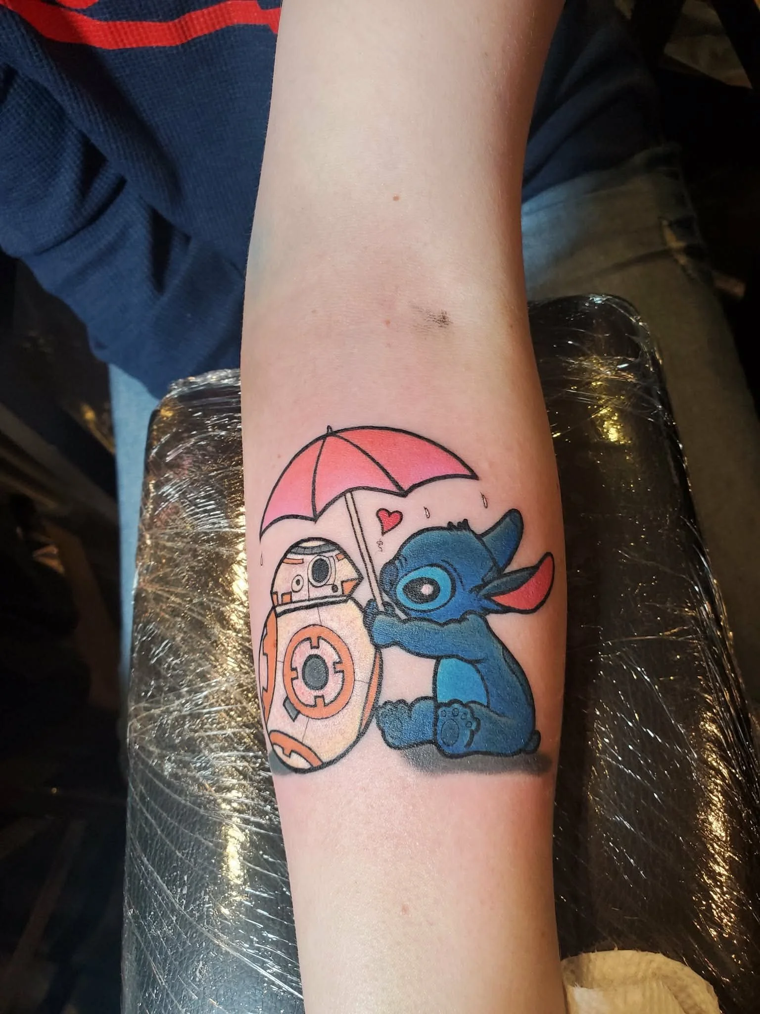 Tattoo of Stitch from Lilo & Stitch holding a pink umbrella with a small red heart above, along with a round droid-like character, on a person's forearm.