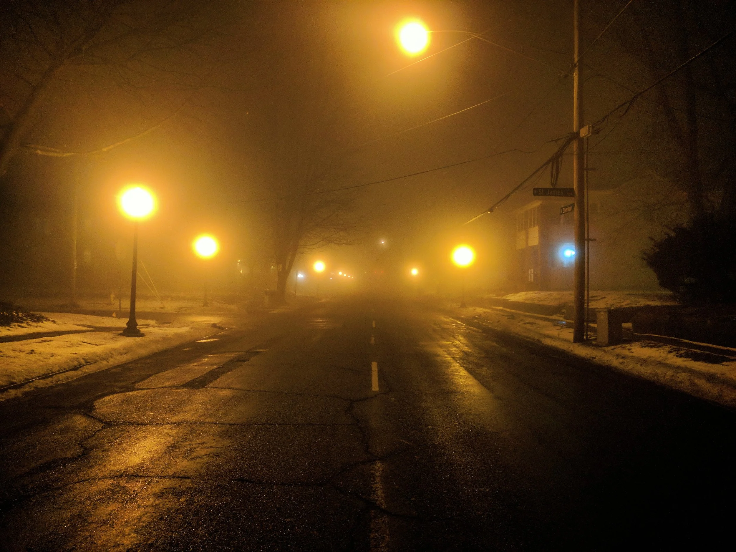 A foggy street scene at night illuminated by several yellow street lights, with snow on the sides of the dark, wet road and a street sign reading 'St. James'.