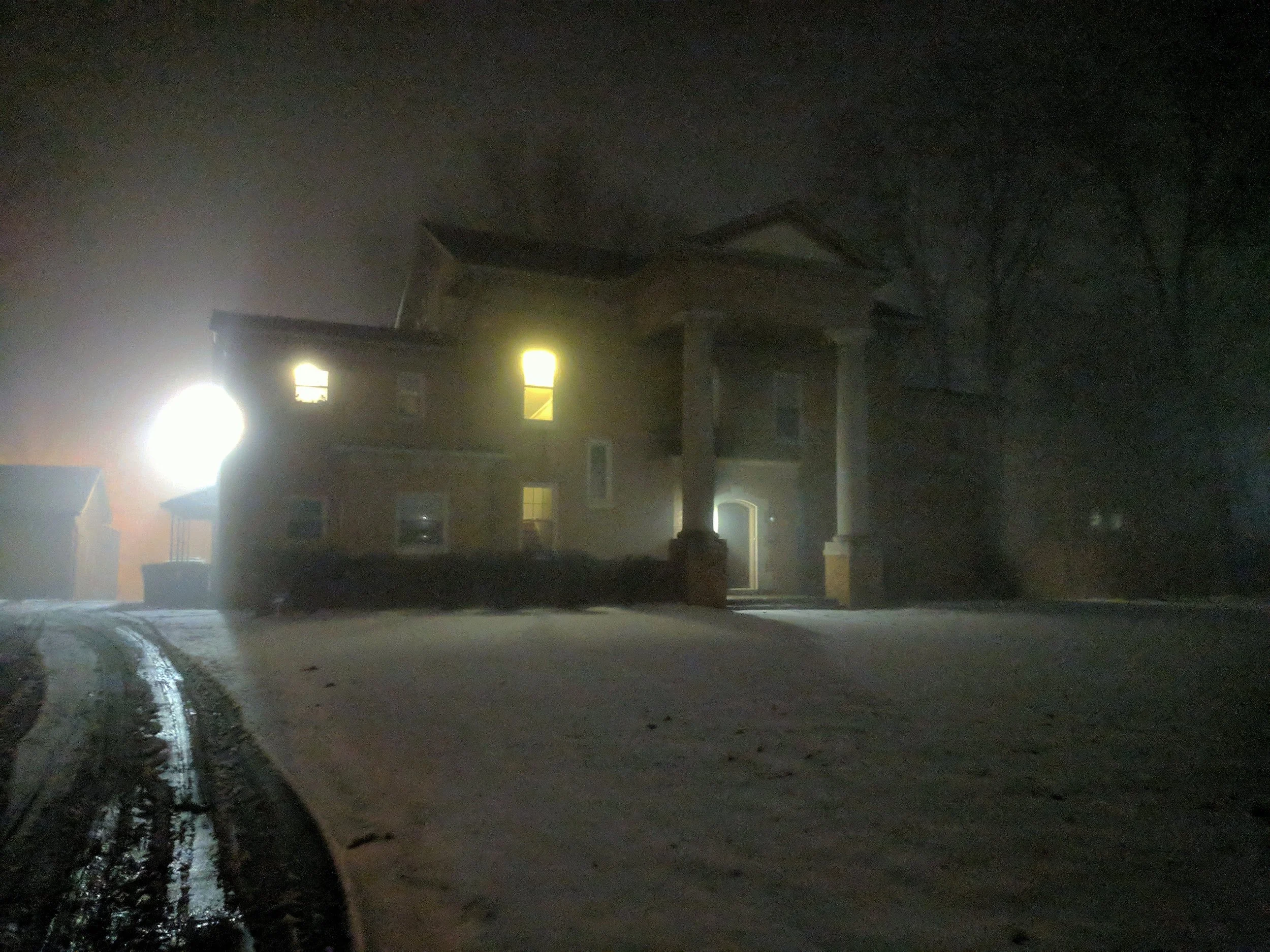 A large house on a snowy night with lights on inside, illuminated by a bright exterior light, and tire tracks in the snow leading up to the house.