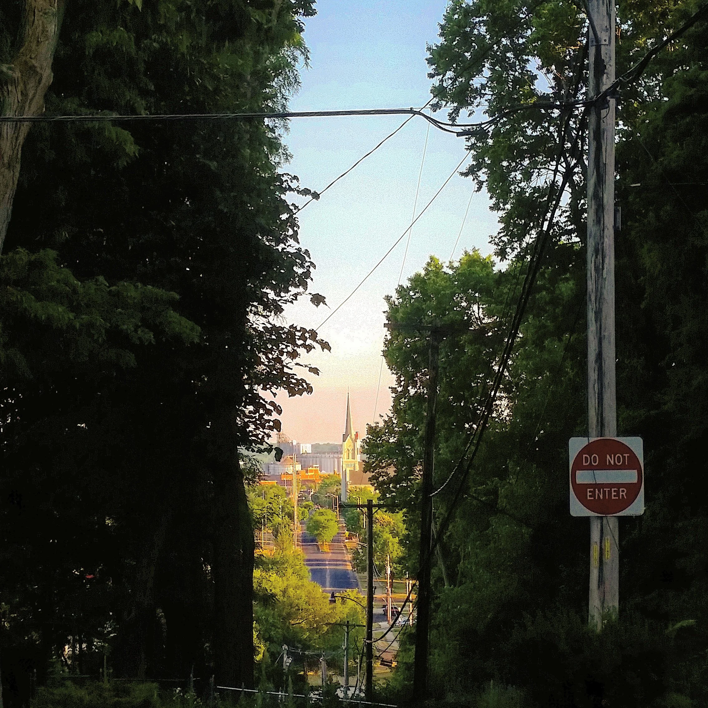 A street view with trees on both sides, utility poles, and wires, leading to a cityscape with a church steeple in the distance. A 'Do Not Enter' sign is visible on the right side of the image.