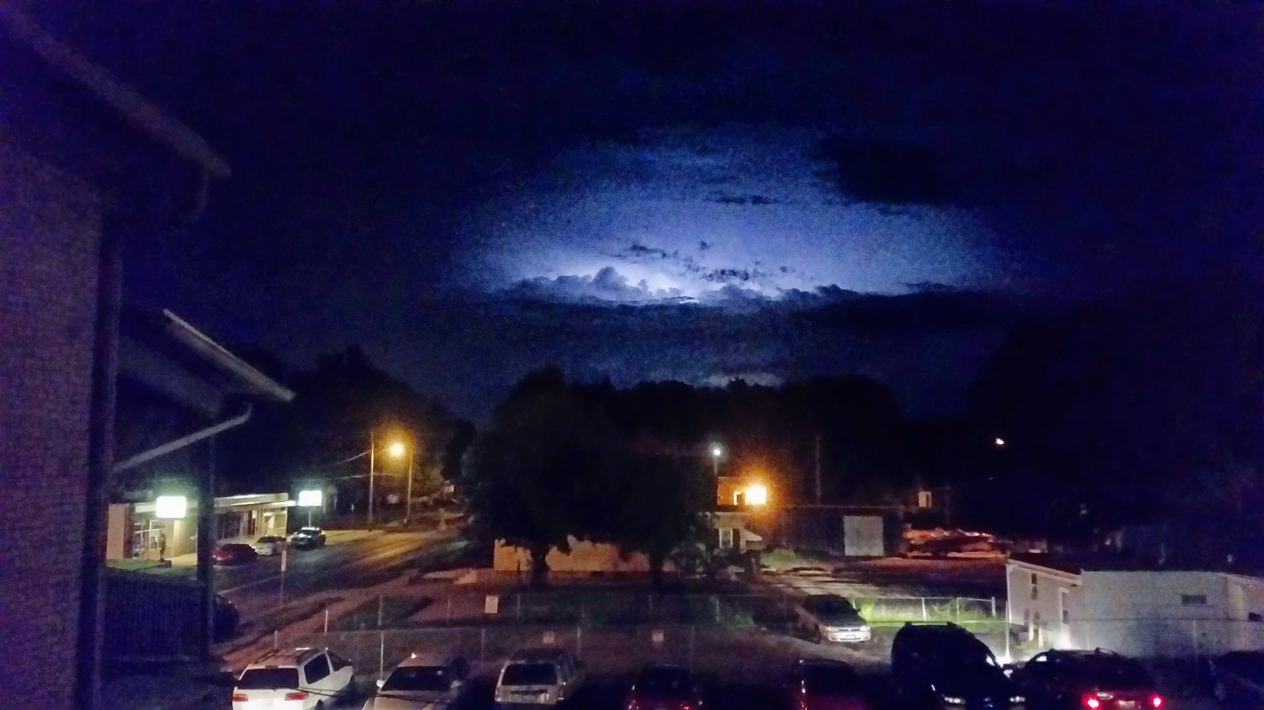 Nighttime view of a parking lot with several cars, a building with lit signs, trees, and streetlights, with dark cloudy sky illuminated by lightning in the background.
