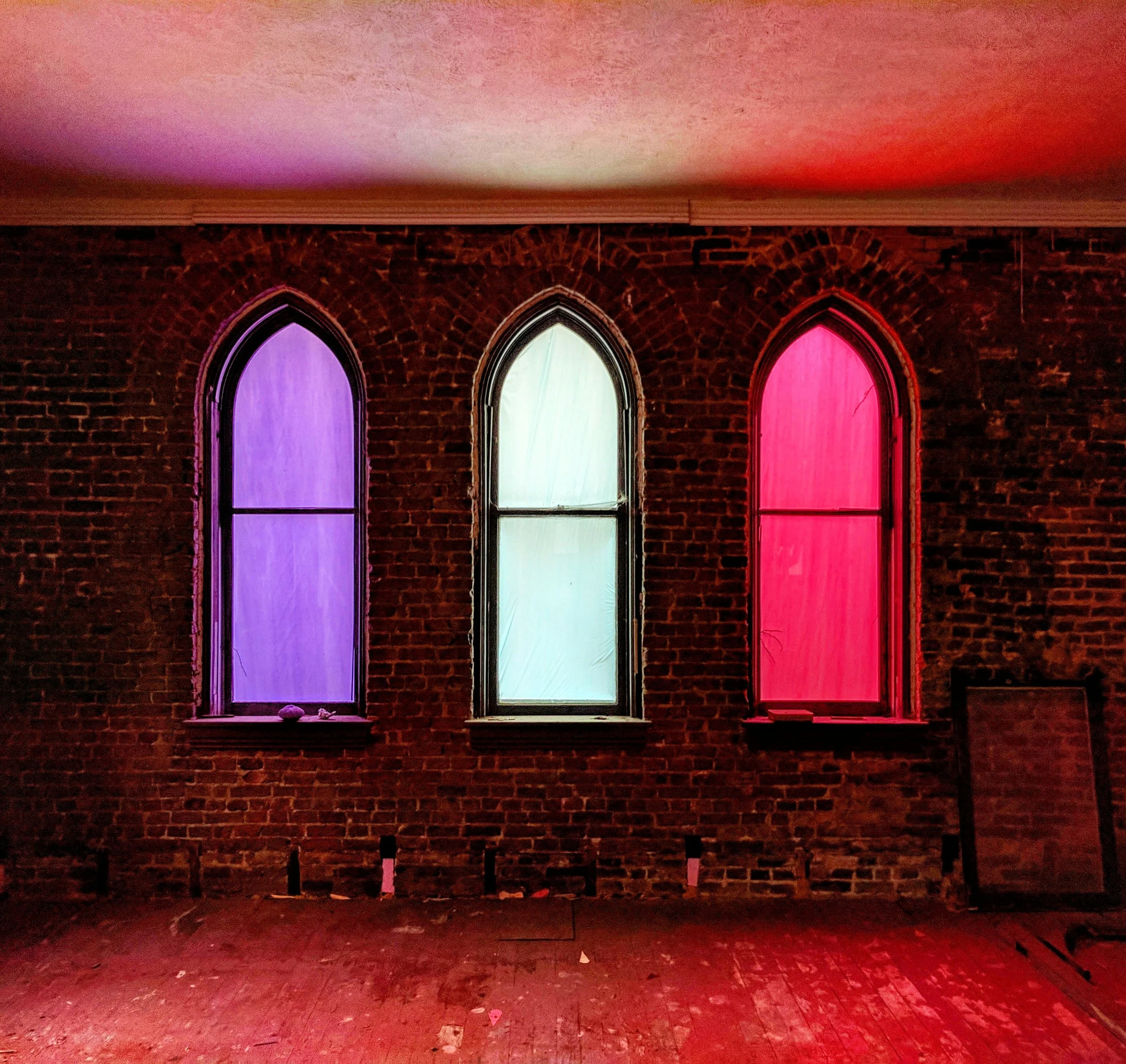 Three arched windows on a brick wall illuminated with purple, white, and red lights.