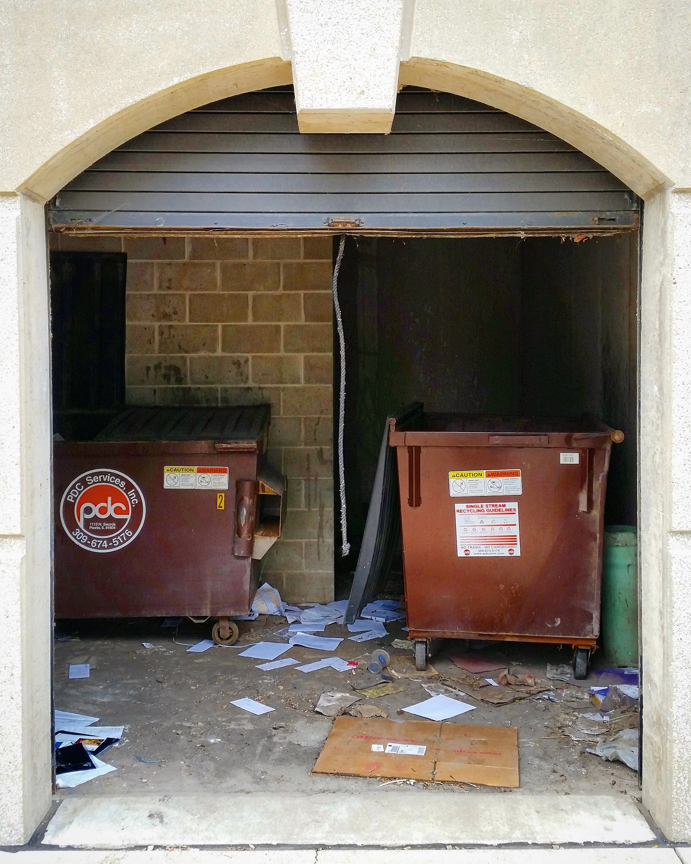 Two overflowing trash bins with scattered papers and debris on the ground inside a garage or storage area.