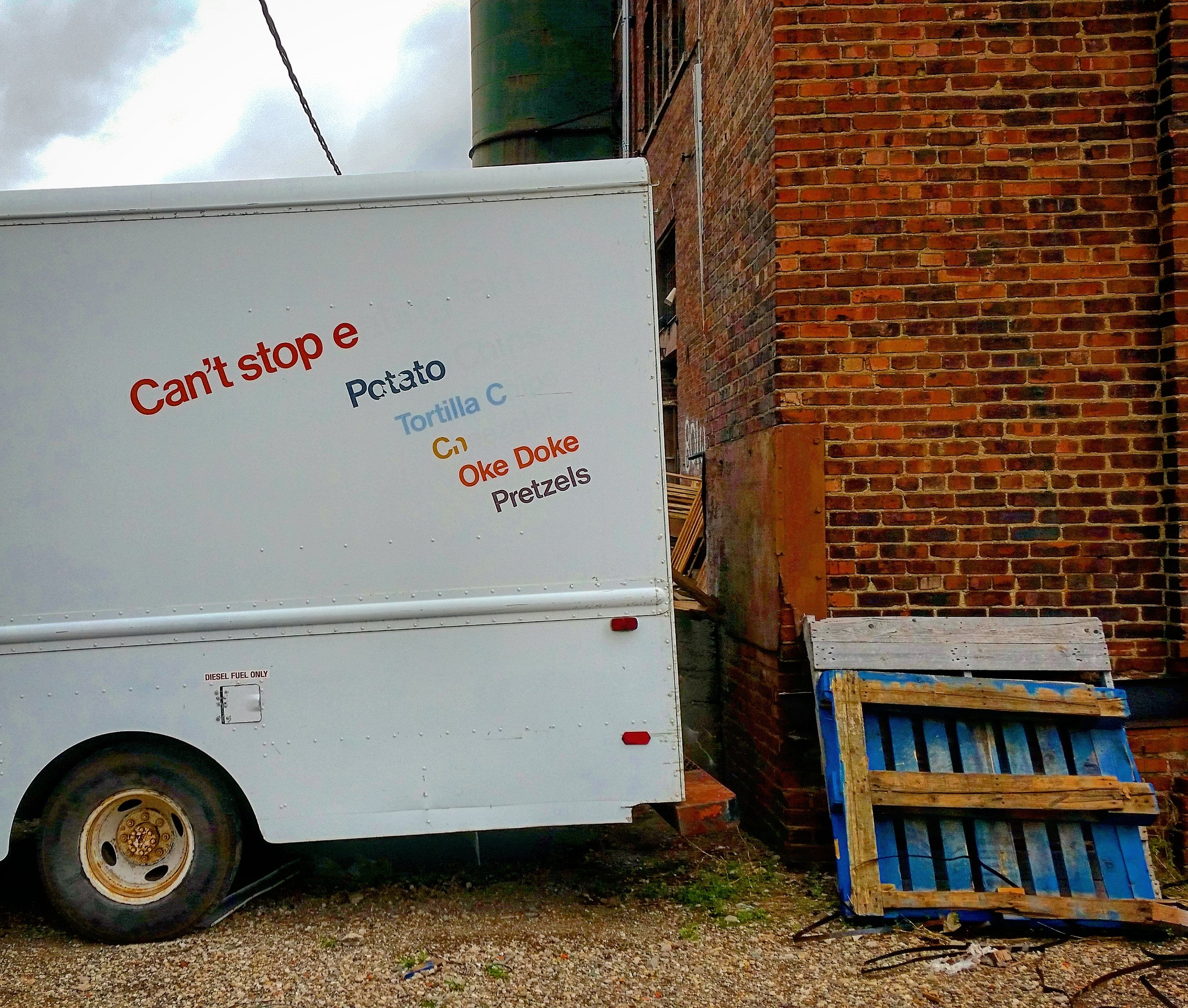 A white food truck with colorful text on the side that reads, "Can't stop e, Potato, Tortilla C, G, Oke Doke, Pretzels." The truck is next to a red brick building, and there is a wooden and blue pallet on the ground nearby.