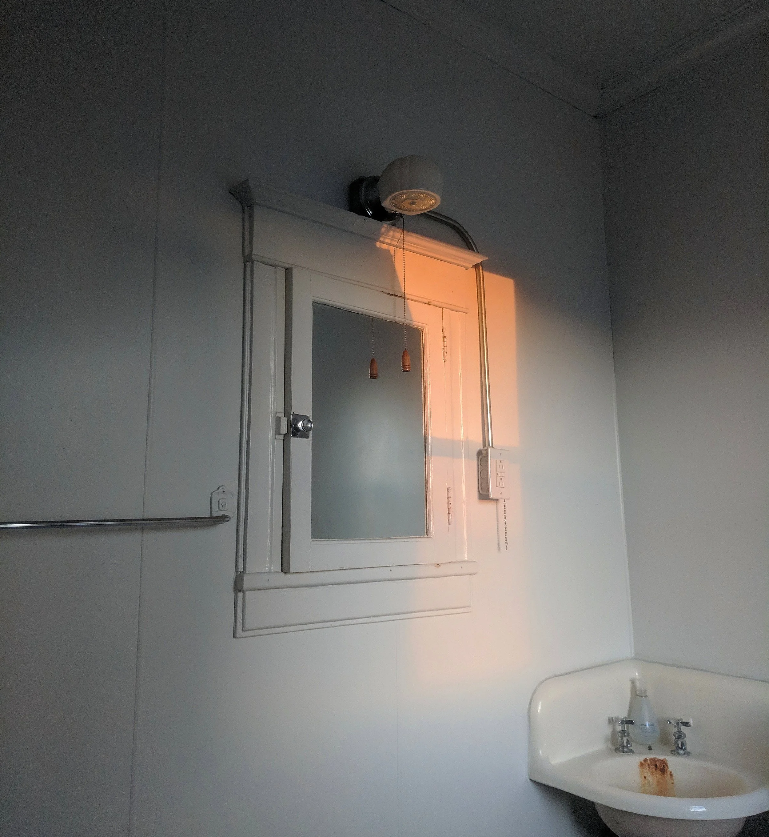 A small wall-mounted sink with rust stain, a mirror with frosted glass, and an old-style light fixtures casting warm orange light on a plain white wall in a room, possibly a bathroom.
