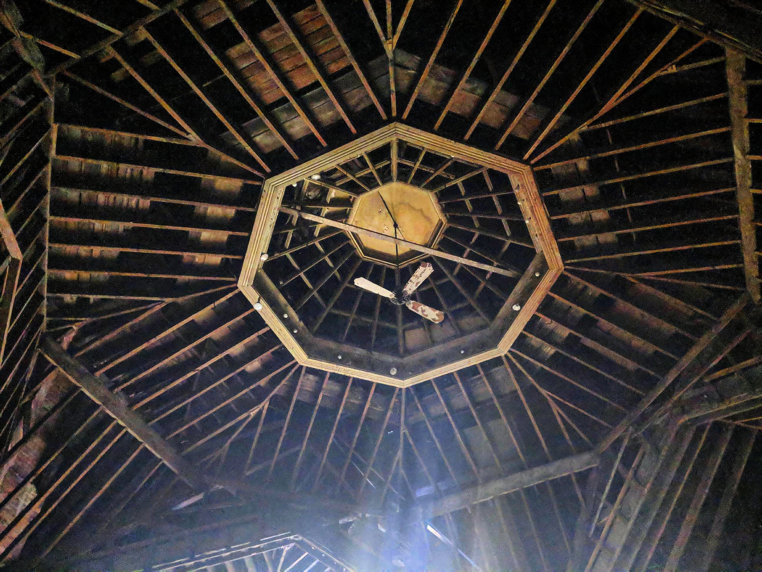 Looking up at a wooden ceiling with a hexagonal shape at the center, featuring ceiling fans and a light fixture.
