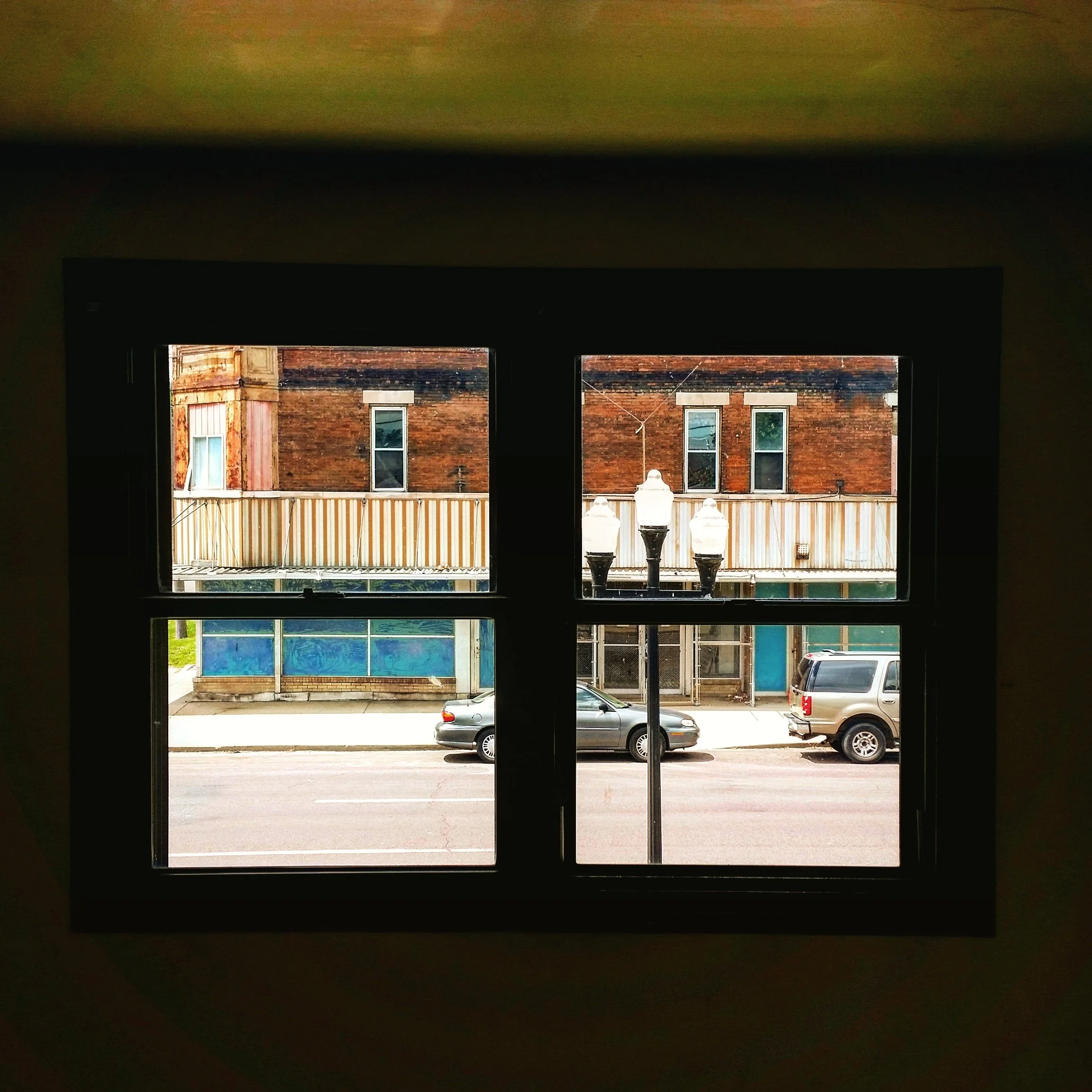 View through a four-pane window showing a brick building with two windows, a street lamp, parked cars, and a sidewalk outside.