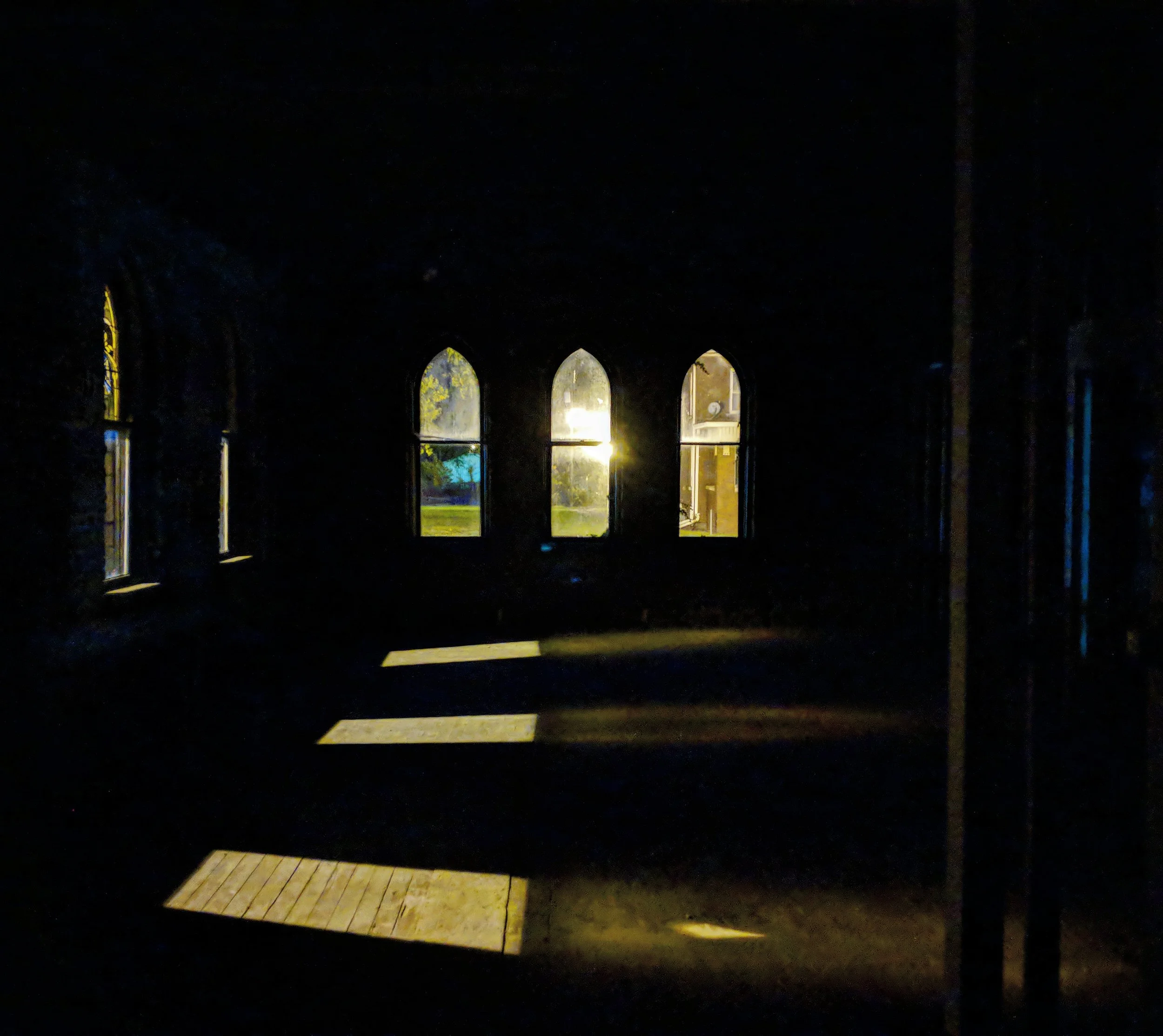 Dark interior with sunlight streaming through four arched windows, casting shadows on wooden floor.