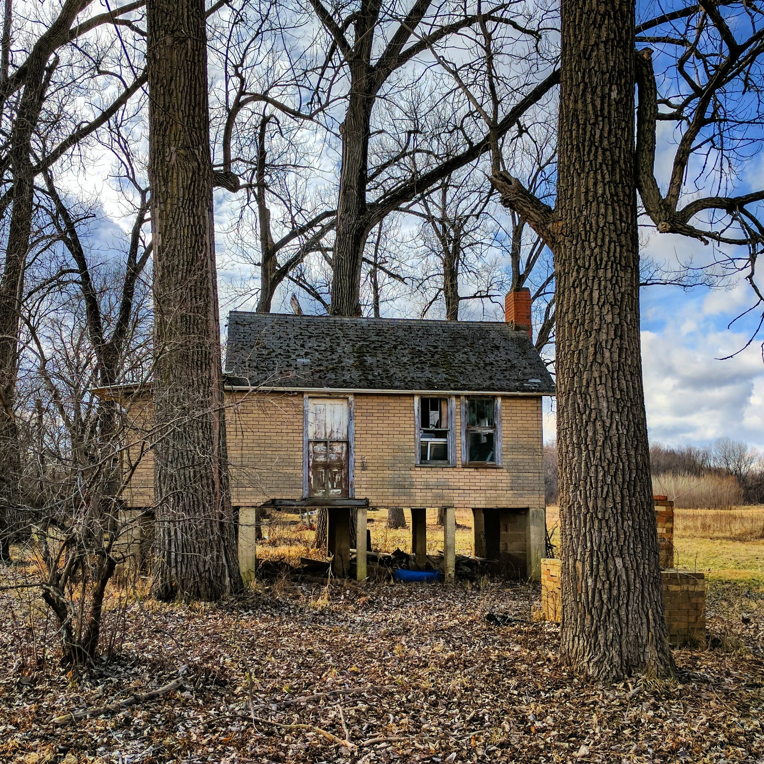 Old house with boarded-up and broken windows, elevated on stilts, in a wooded area with leafless trees and a cloudy sky.