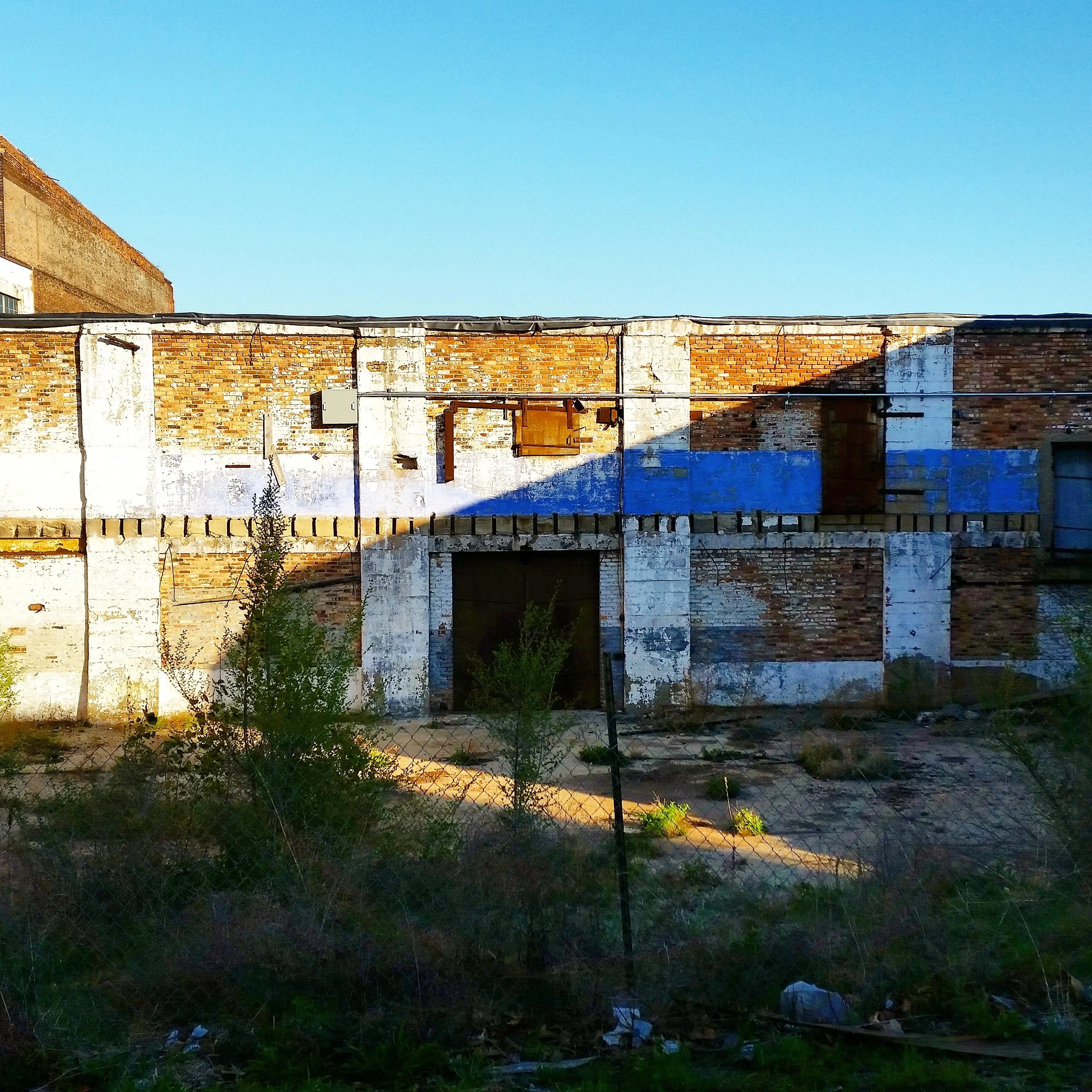 A dilapidated brick building with some painted and some exposed bricks, a boarded-up window, and a shadow cast across the front. There is a small patch of overgrown vegetation and a wire fence in the foreground, with trash scattered on the ground.