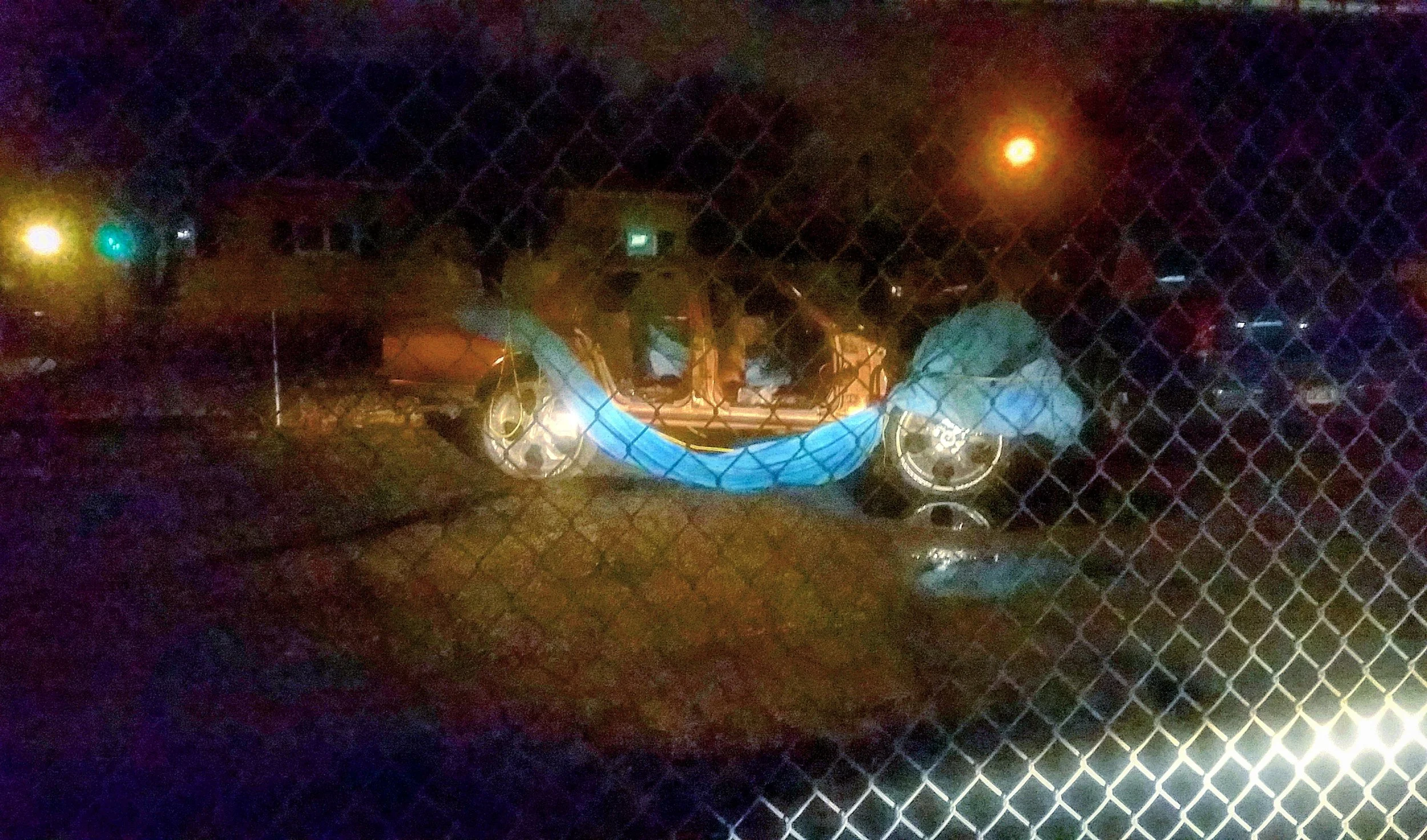 A damaged orange car with parts disassembled, covered with a blue tarp, seen through a chain-link fence at night.