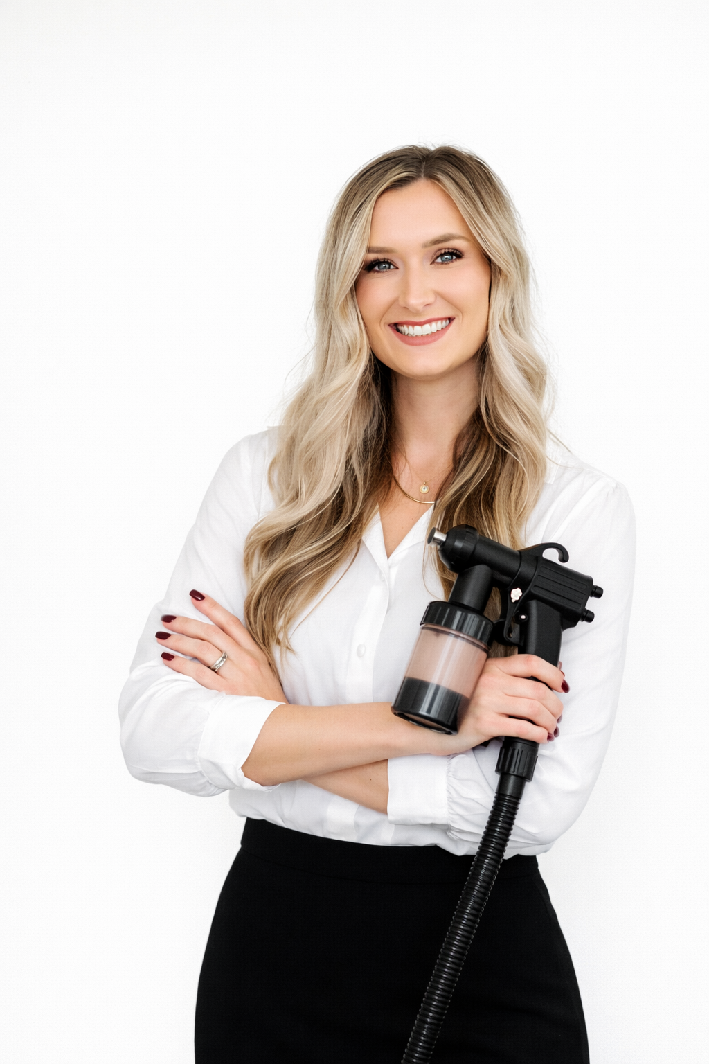 A woman with long blonde hair, wearing a white blouse and black pants, smiling and holding a spray gun with a container, against a plain white background.