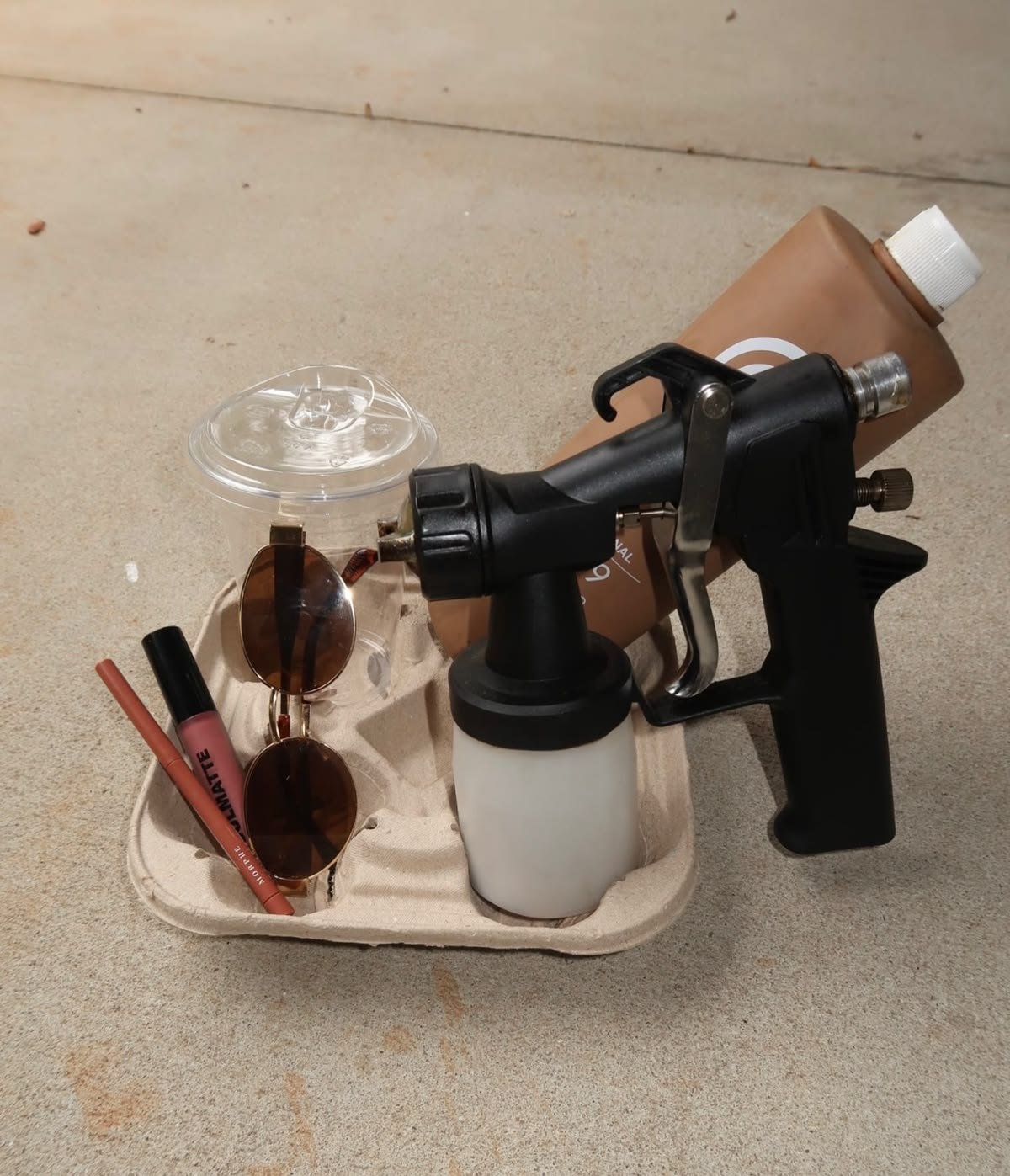 Cup holder holding a spray tan gun, makeup, sunglasses, and spray tan solution.