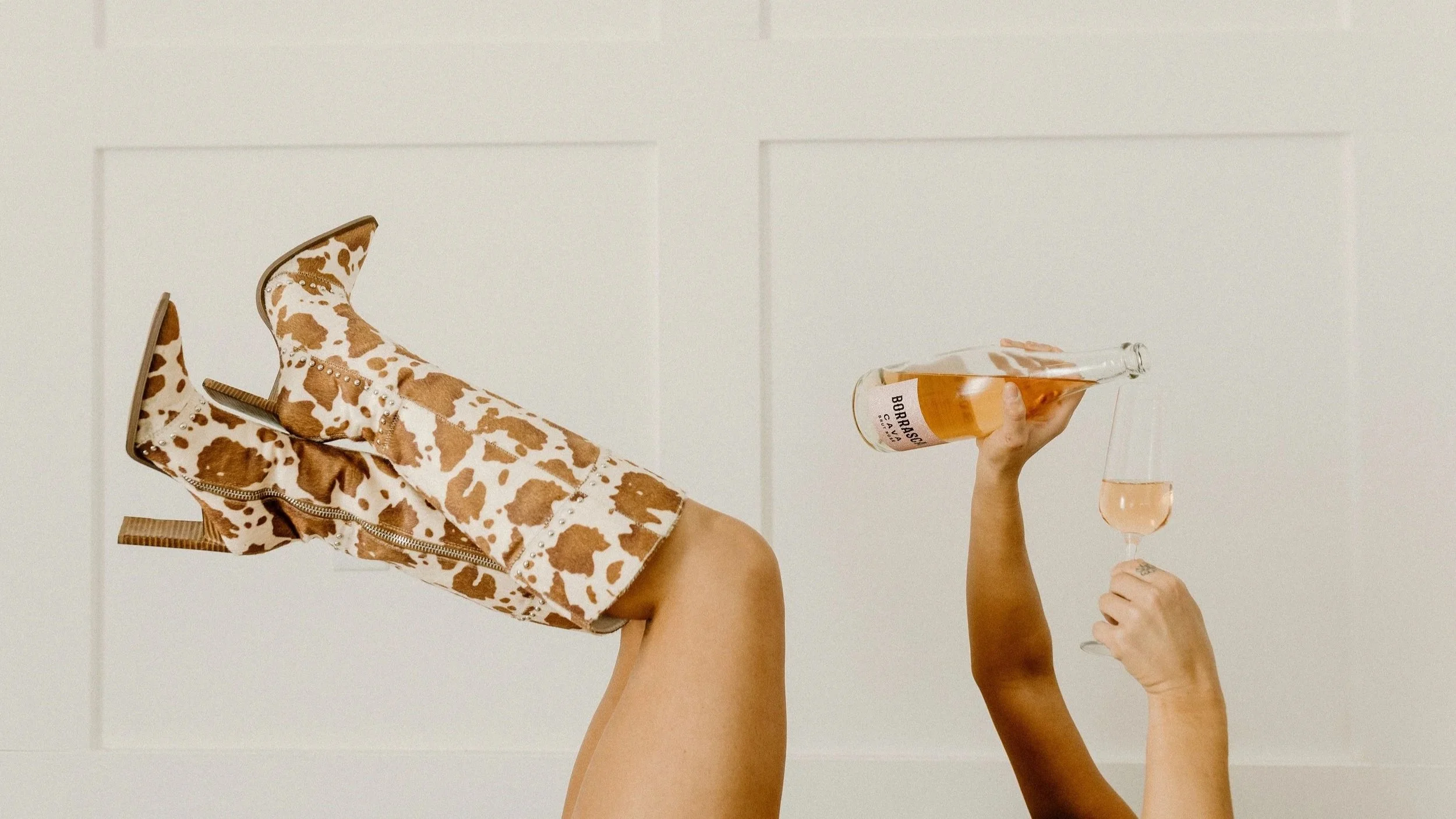 Person lying with legs raised, wearing cowboy boots with a cow print pattern and zippers. They hold a bottle of rosé wine and pour it into a glass.