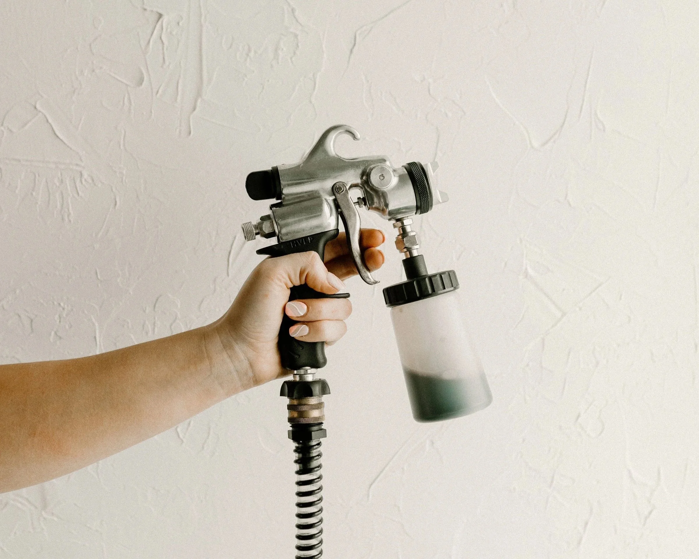 Hand holding a spray gun with a container of paint or fluid, against a white textured wall.