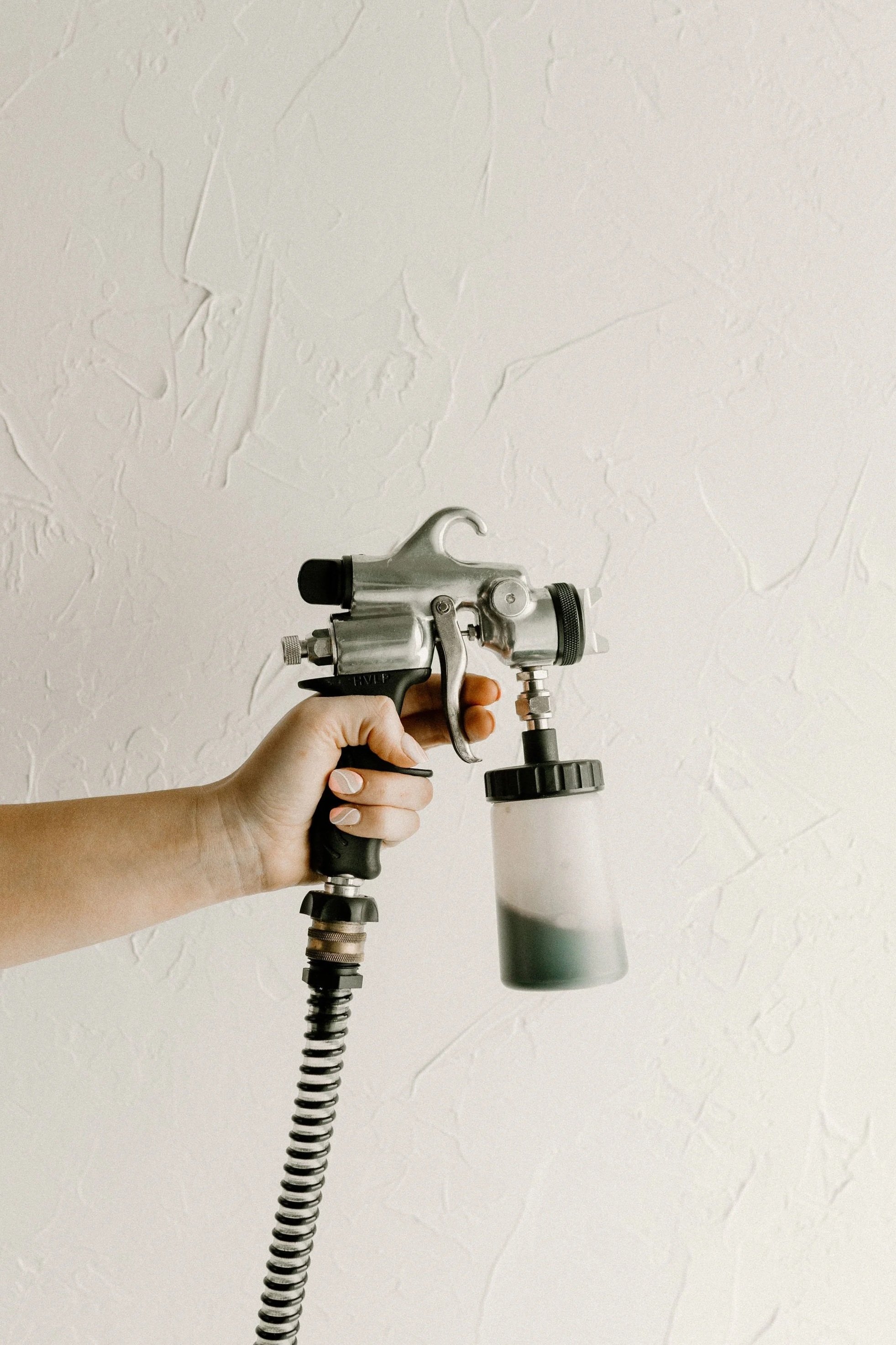 A person holding a spray gun with a container attached, against a plain textured wall.