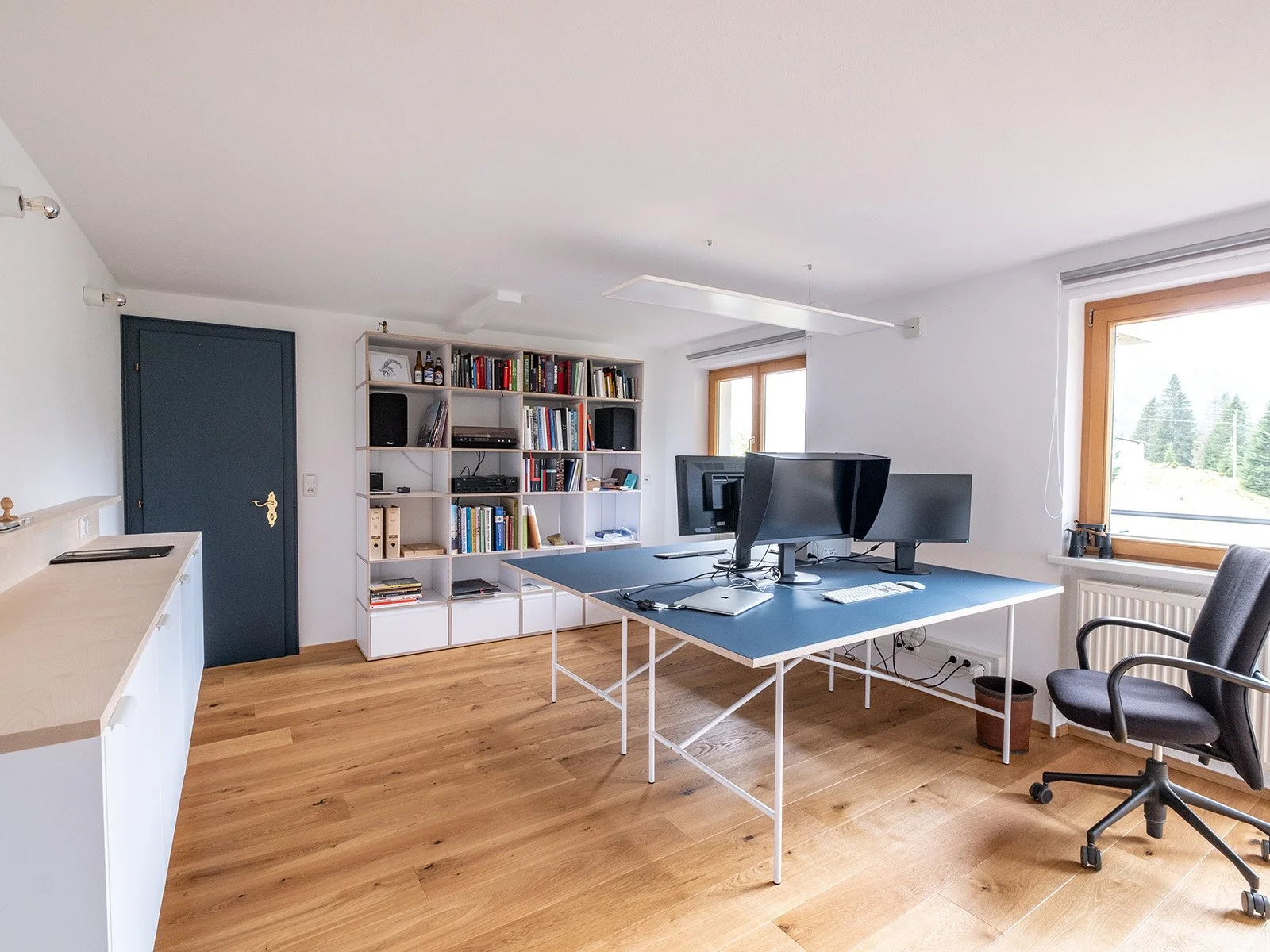 Arbeitszimmer mit Schreibtisch, mehreren Computern, einem Bücherregal, Stuhl und Fenster mit Sicht auf Bäume und Himmel.