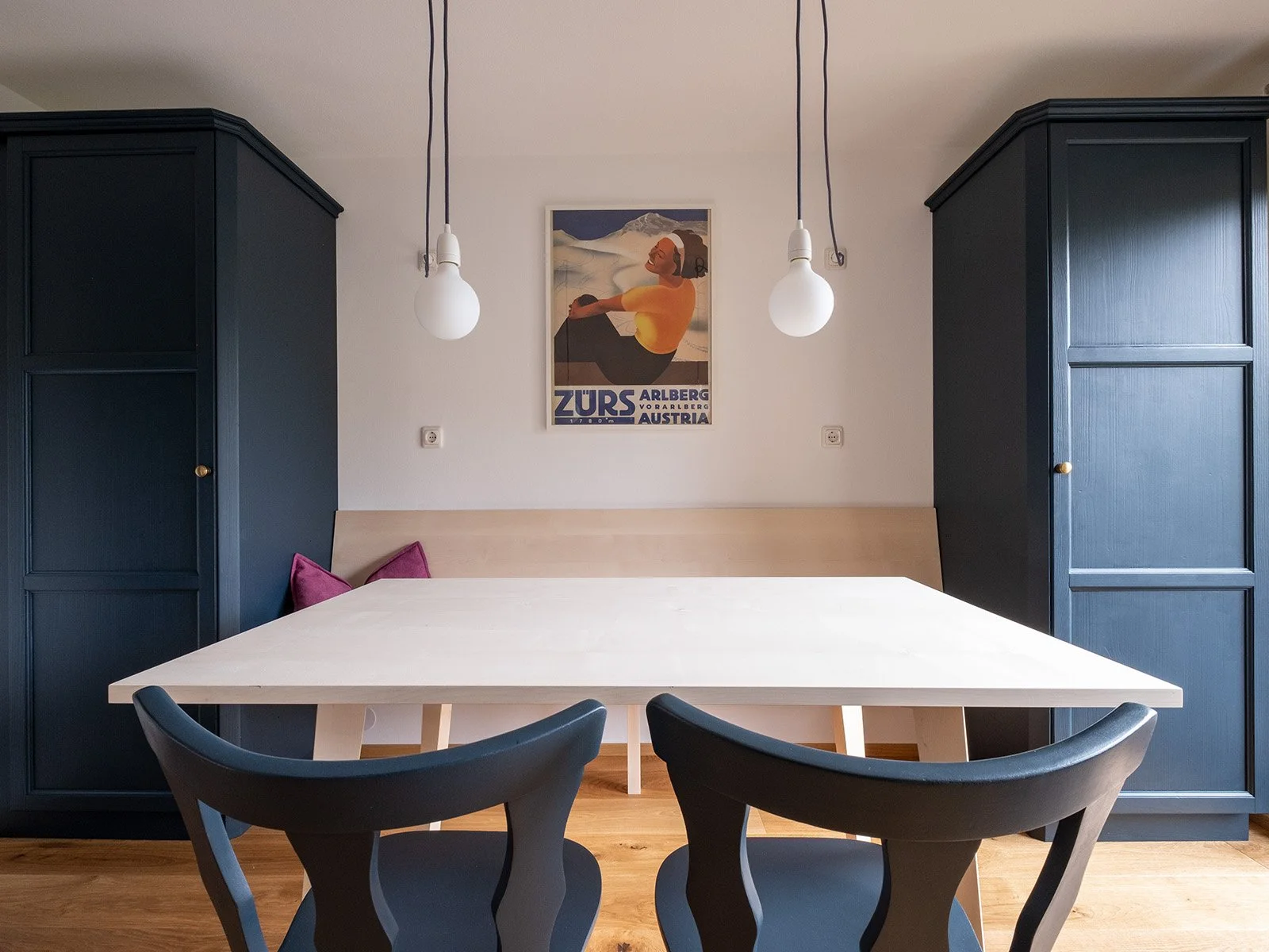 Ein Tisch im Esszimmer mit zwei schwarzen Stühlen, ein beige farbener Hintergrund, über dem Tisch hängen zwei weiße Beleuchtungskörper, an der Wand das Plakat »Zürs Arlberg Austria«, im Hintergrund stehen zwei dunkelblaue Schränke.