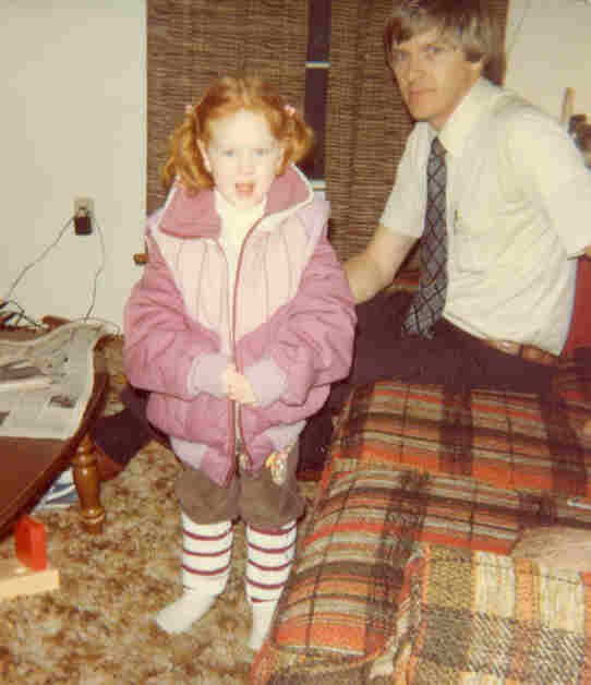 A vintage photo of a young girl with red hair wearing a pink jacket, brown skirt, and striped socks standing next to a man dressed in a white shirt and tie, sitting on a plaid patterned couch in a living room.