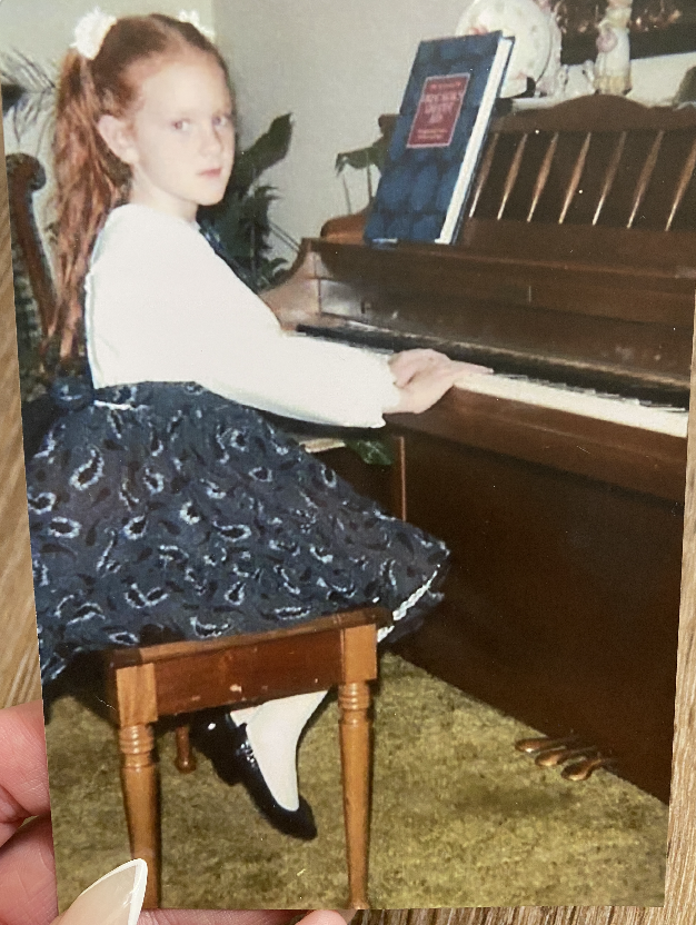 Vintage photograph of a young girl with long red hair in pigtails wearing a black and white dress and black dress shoes sitting at a piano