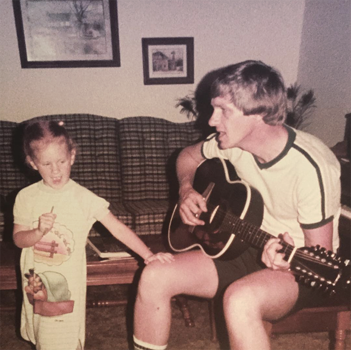 A vintage photograph including a young girl singing with a man playing a guitar in a living room next to a plaid couch