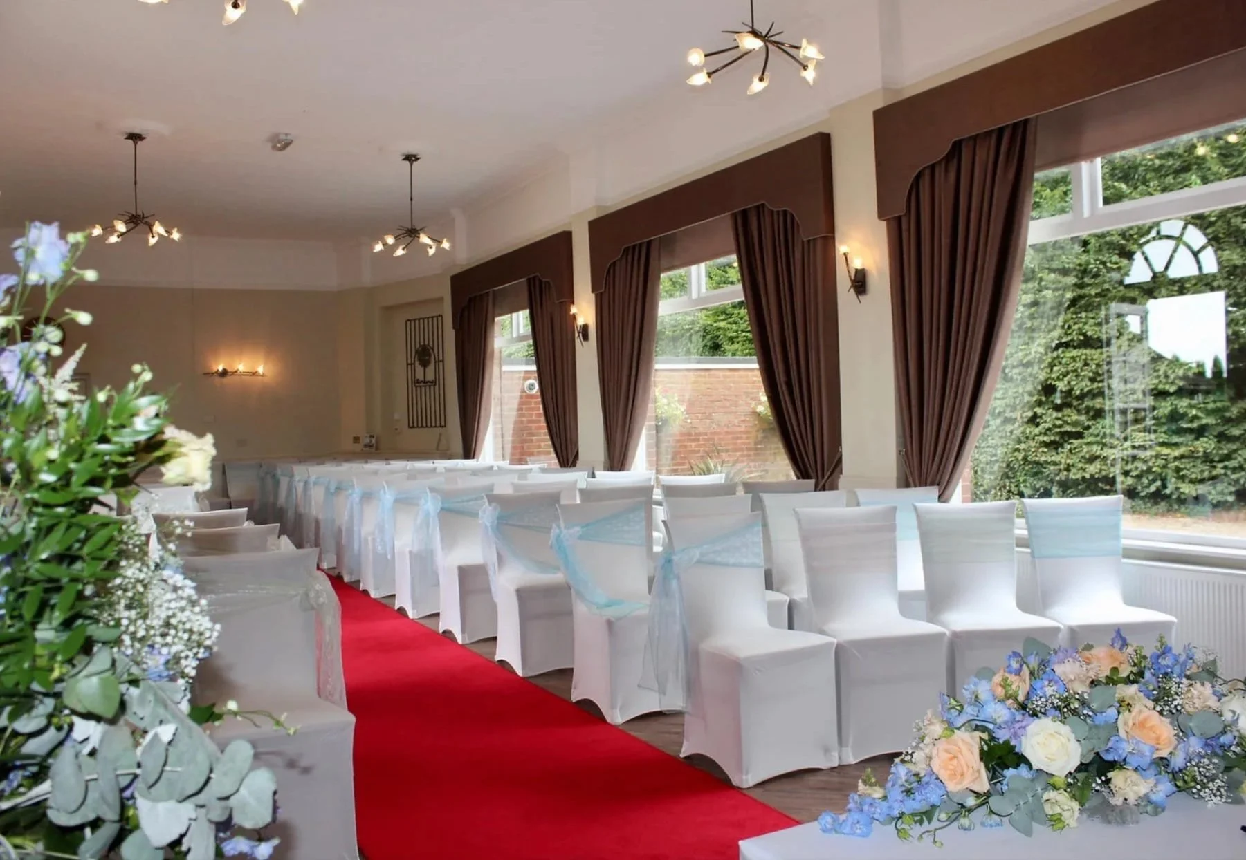 Interior of a decorated event space with white chairs, a red aisle runner, floral arrangements, large windows with brown curtains, and ceiling lights.