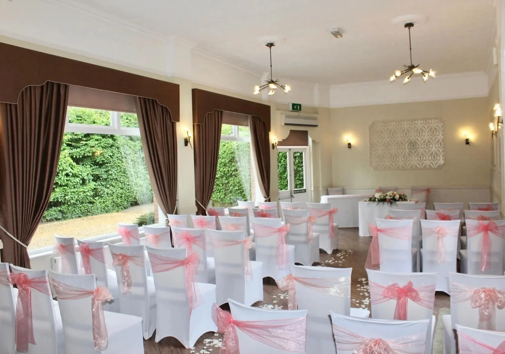 A decorated wedding ceremony room with white chairs adorned with pink ribbons, large windows with brown curtains, and greenery outside. The room has wall-mounted lights, ceiling fixtures, and a floral arrangement on a table at the front.