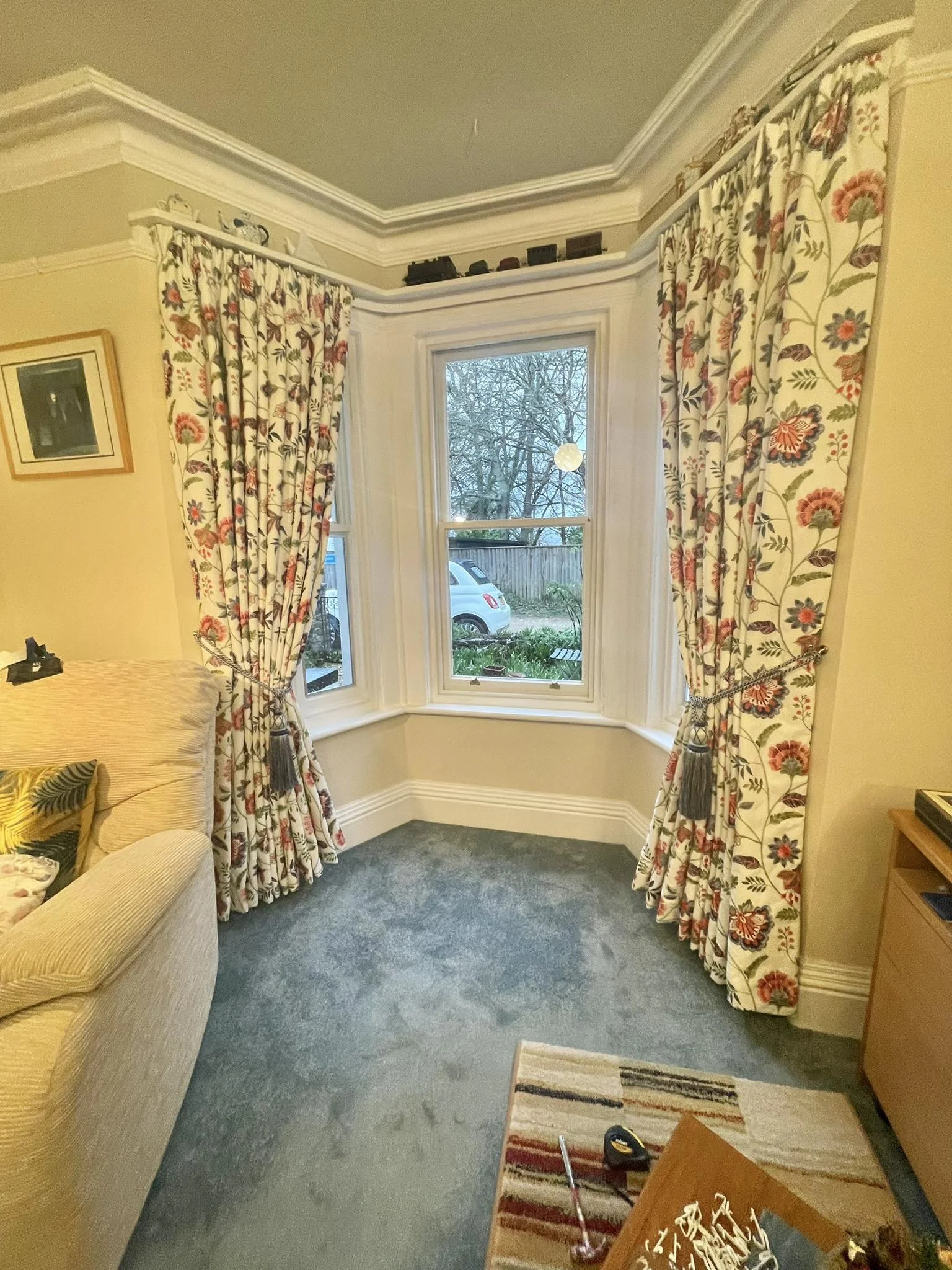 View of a living room corner with a window, floral curtains, a beige sofa, a small wooden table, and a colorful rug on a gray carpeted floor.