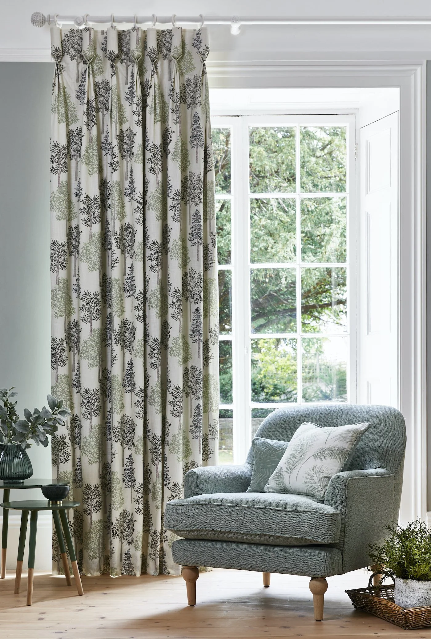 Light blue armchair with a decorative pillow, placed in front of a large window with white frame; a potted plant in a woven basket on the floor next to it; a small side table with a plant and decorative items; floral curtains and greenery outside the window.