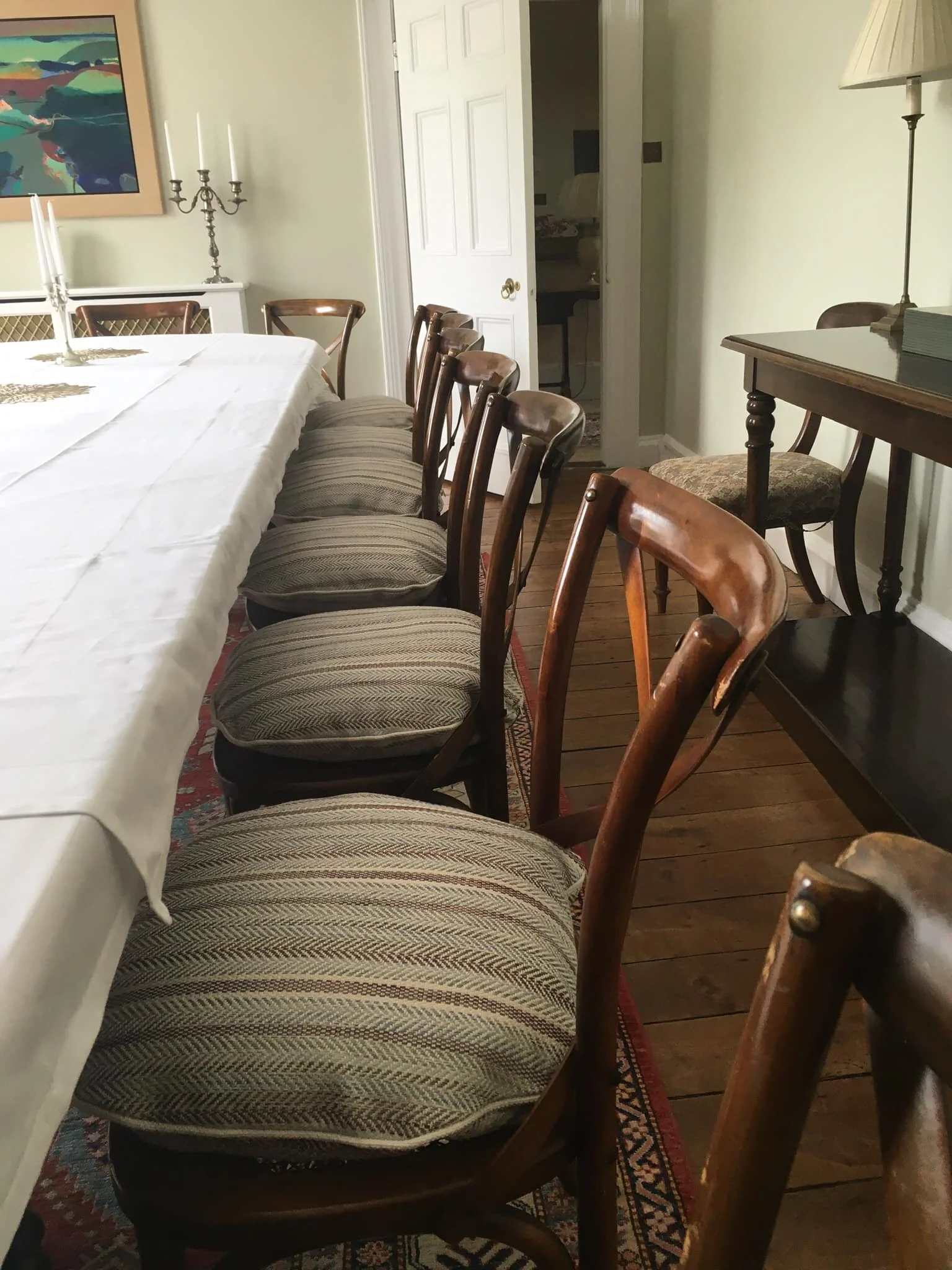 A dining room with a long table covered with a white tablecloth, surrounded by wooden chairs with upholstered seats, a framed colorful landscape painting on the wall, a brass candelabrum with white candles, a wooden side table with a table lamp, and 
