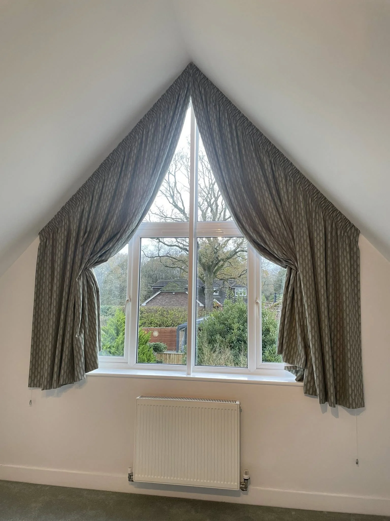 A window with gray patterned curtains tied back, revealing a view of trees and houses outside on a cloudy day. A radiator is located below the window.