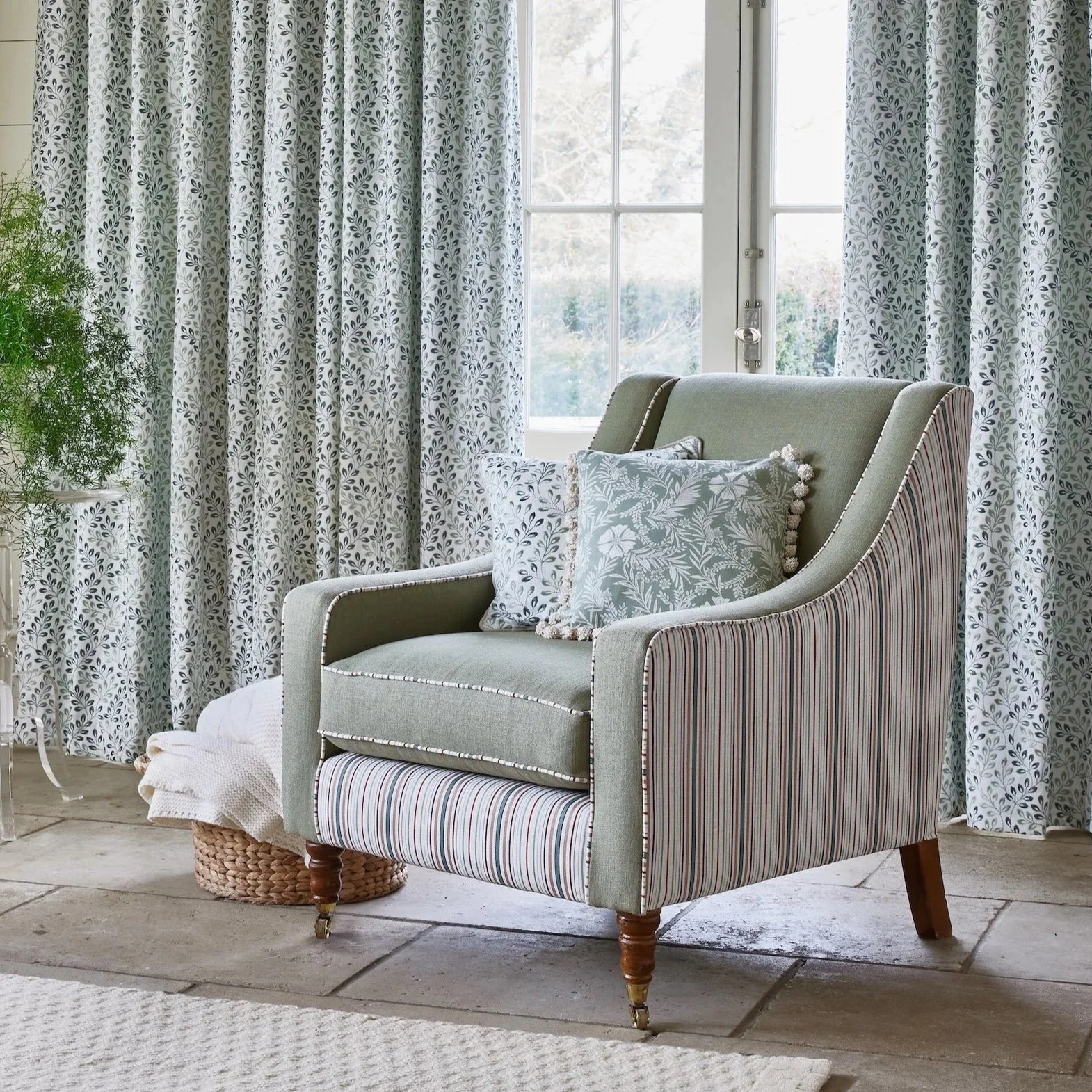 A cozy living room corner with an armchair and decorative pillows, with a large window and patterned curtains in the background, and a potted green plant on the side.
