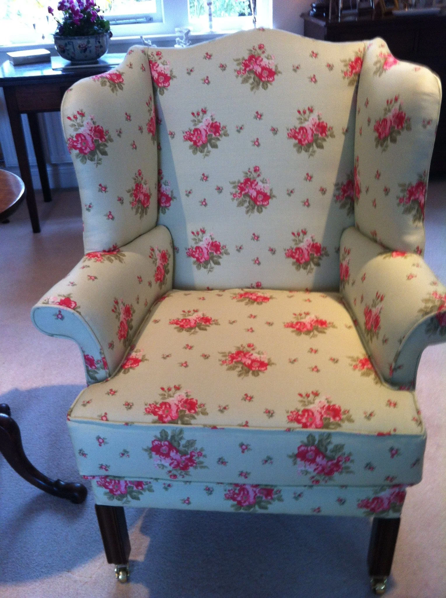 A vintage armchair with a floral pattern of pink and red roses on a cream background. The chair has rounded arms and a high back, situated in a cozy room with a window, a wooden side table with a potted plant, and various decorative items.
