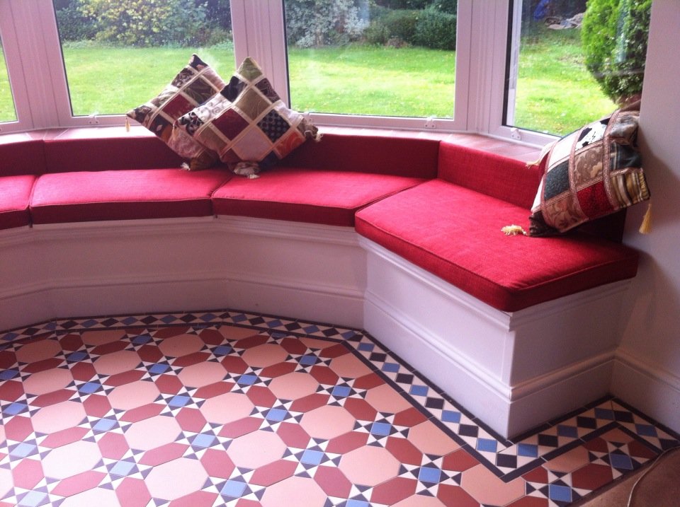 Built-in window seat with red cushions and decorative plaid pillows in a sunroom with large windows and geometric patterned tile floor.