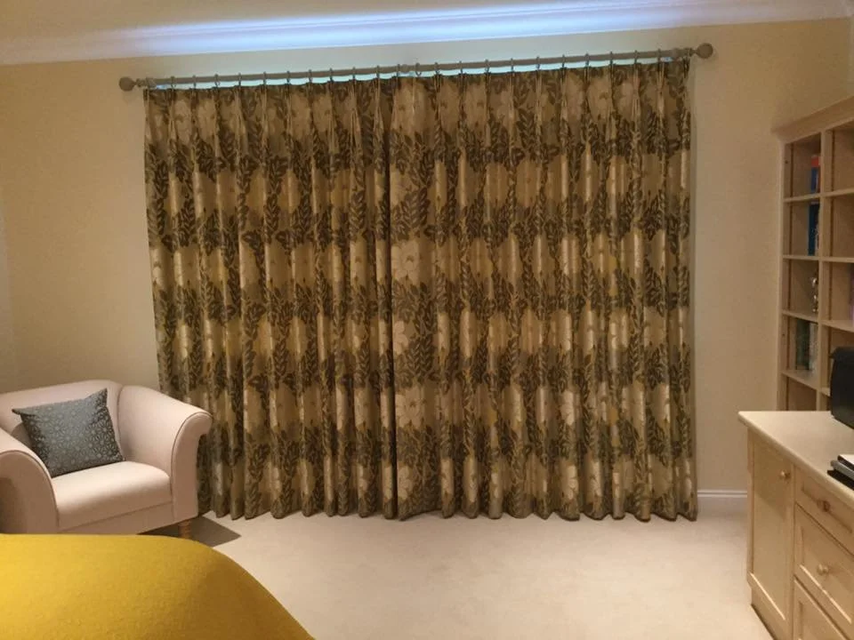 A living room with a beige sofa and a patterned pillow on it, covered by closed floral curtains, with a wooden bookcase on the right and a piece of furniture with a computer or speaker on it, next to the bookcase.