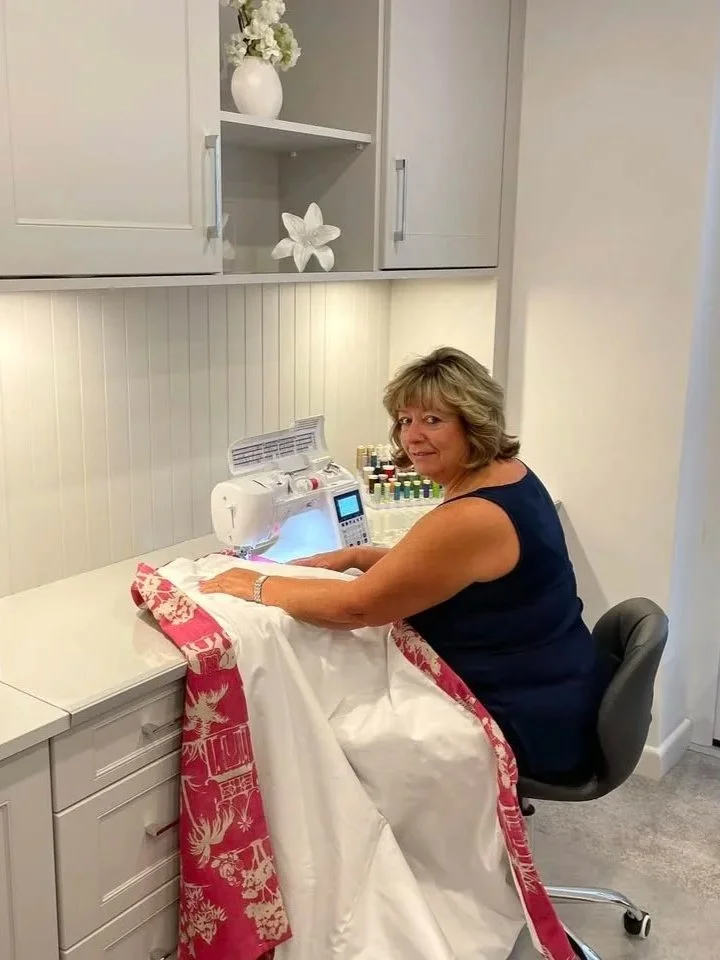 Pauline is sitting at a sewing machine in a craft room, surrounded by rolled fabric and bottles of nail polish, with white cabinets and decorative items in the background.