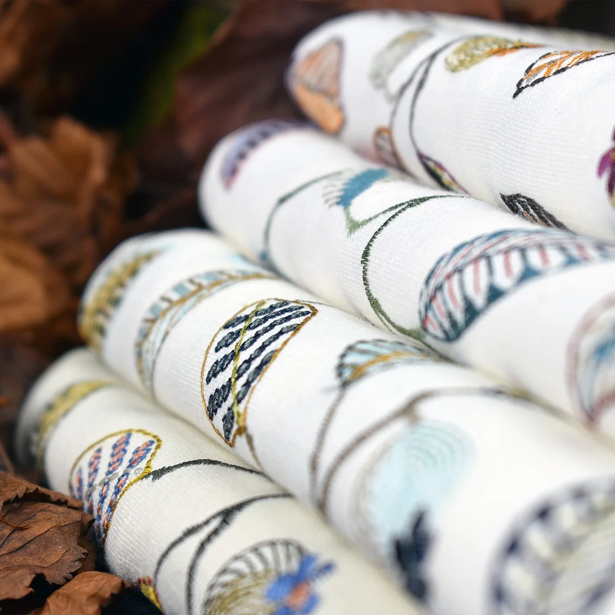 Close-up of rolled white fabric with colorful embroidery of eggs and leaves, lying on brown leaves.