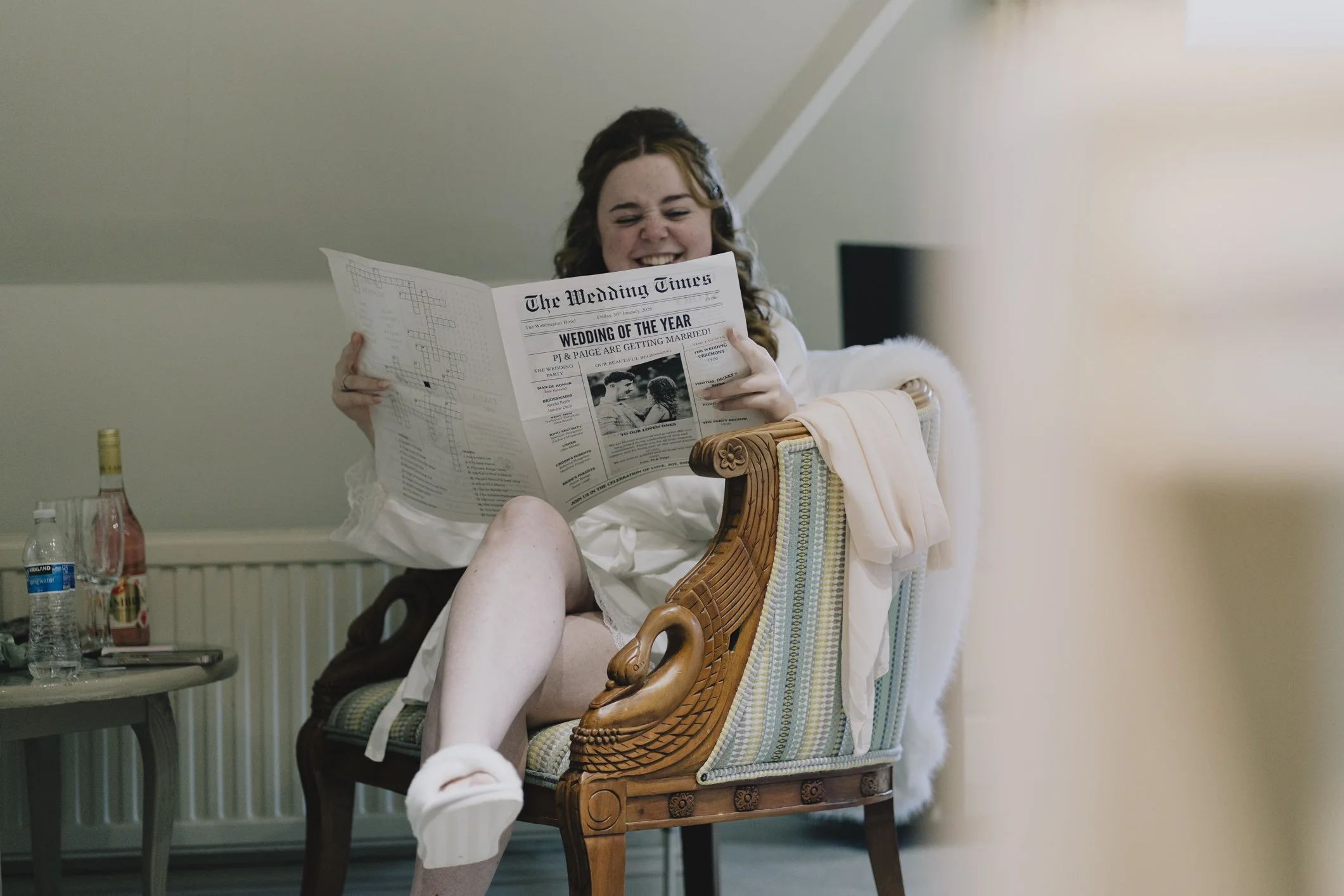 Bride in a white robe sitting on a decorated wooden armchair, reading a newspaper titled 'The Wedding Times' with a headline 'Wedding of the Year'.