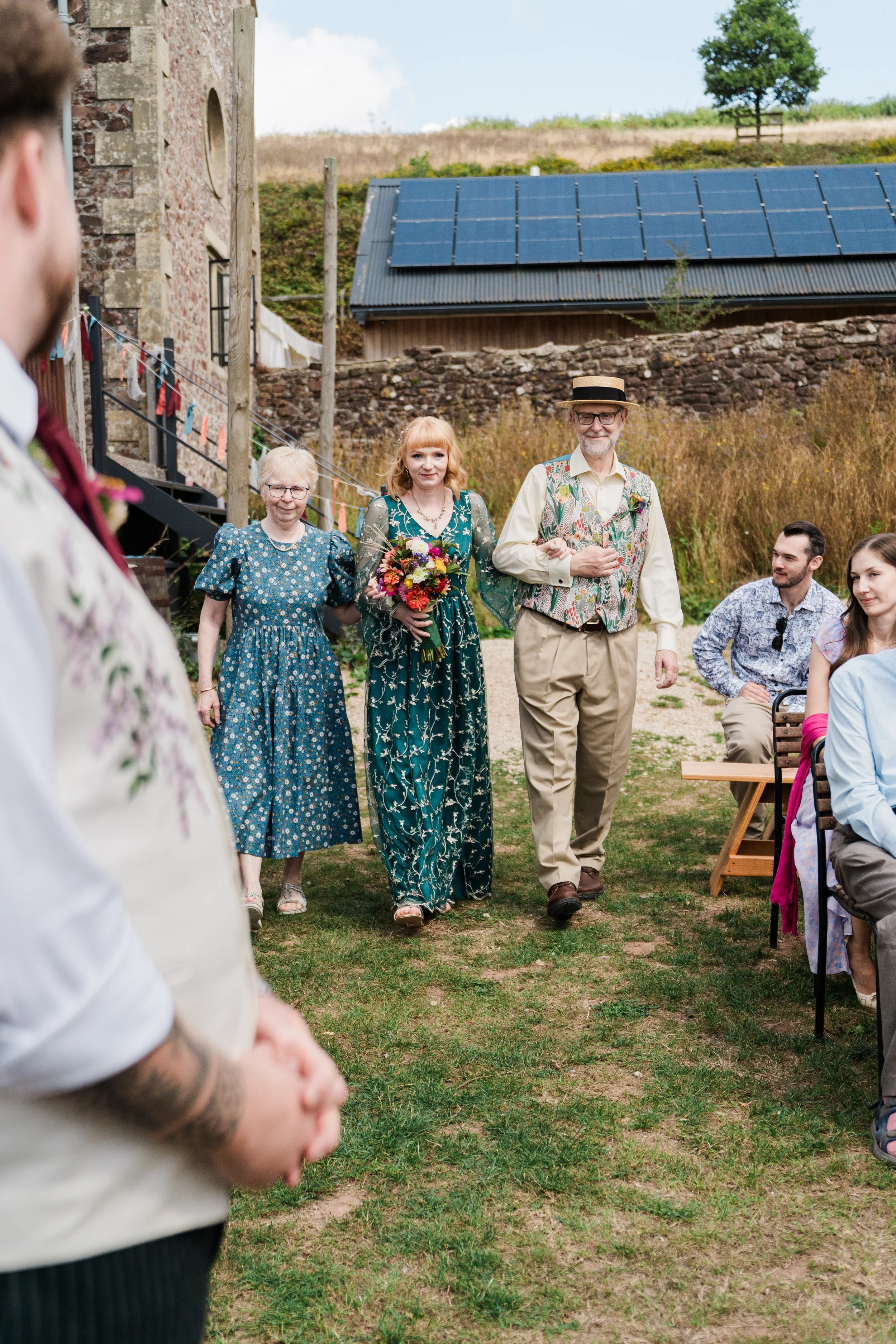 Group of people gathered outdoors during a celebration, with some walking and others sitting, in a rustic setting with a stone building, a barn with solar panels, and a tree in the background.