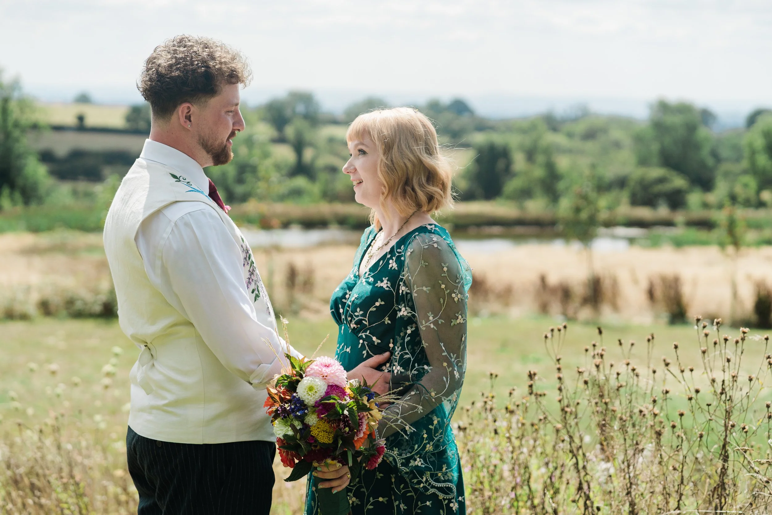 A couple standing close together outdoors, holding a colorful bouquet of flowers at Lapwing Farm, with a scenic landscape in the background.