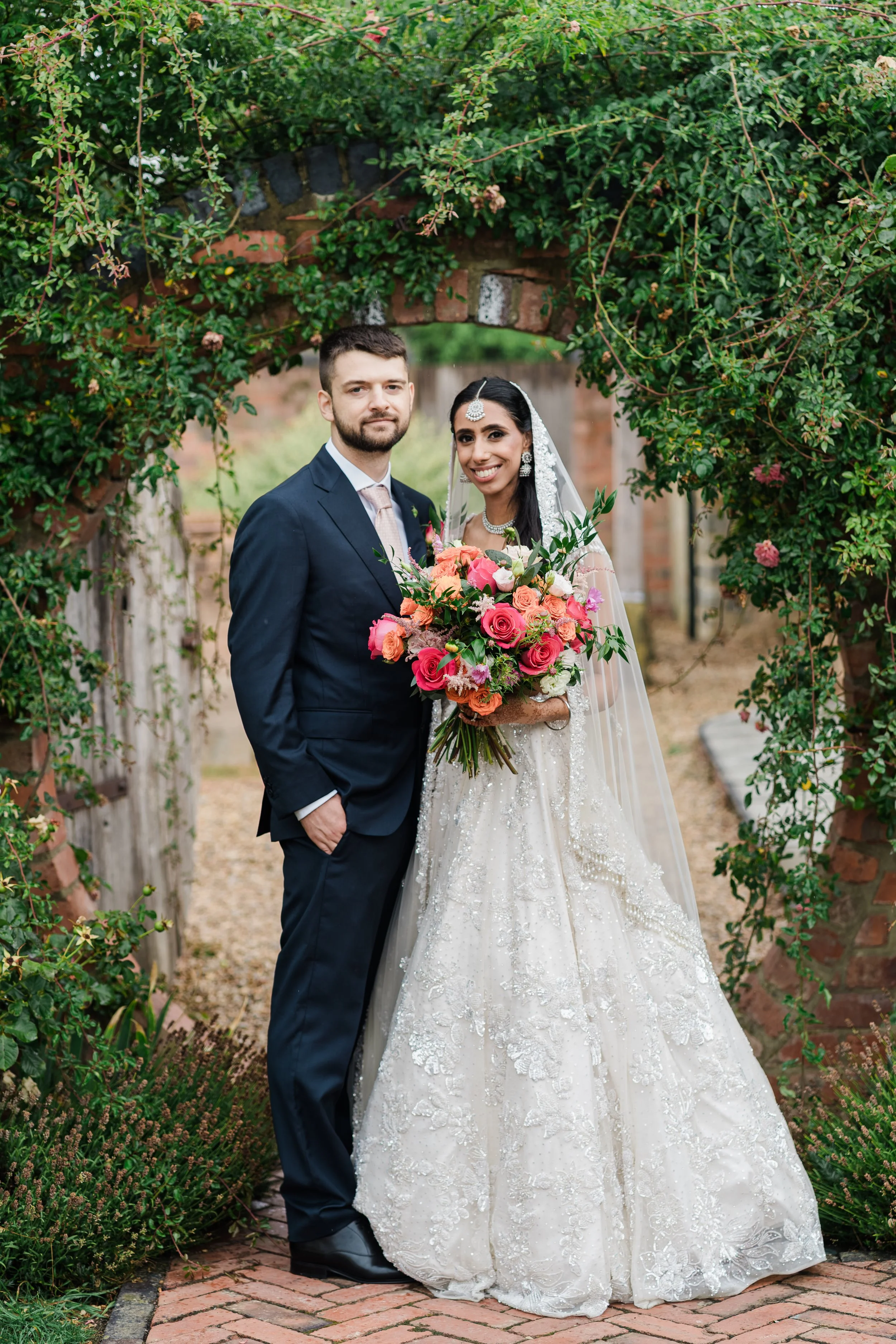 A bride and groom posing under a garden archway decorated with greenery and pink flowers. The bride is holding a bouquet of flowers. The groom is wearing a navy suit with a light pink tie capture by Blackwell Grange Wedding Photographer