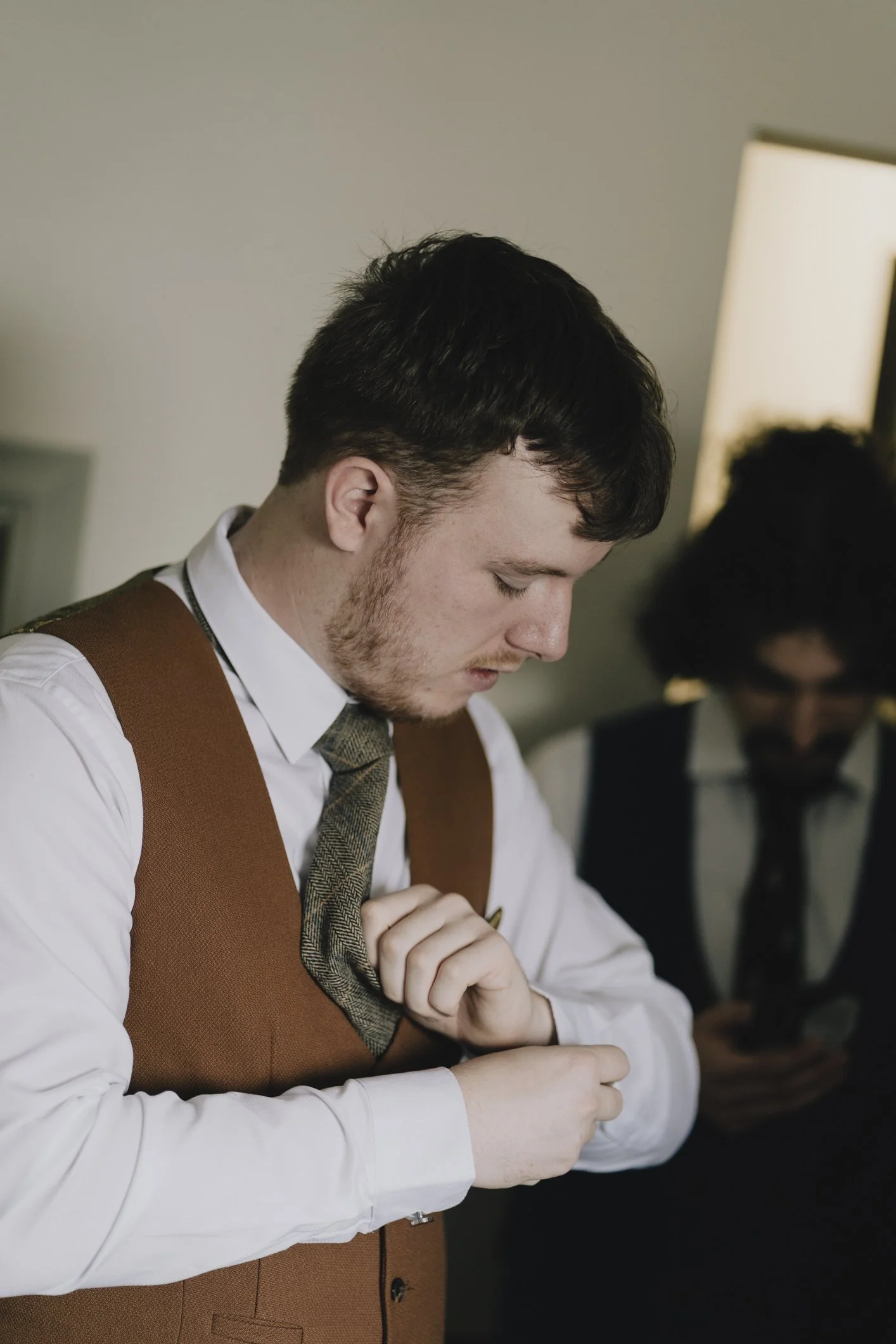 Groom in brown vest adjusting his tie, beside another person using a phone.