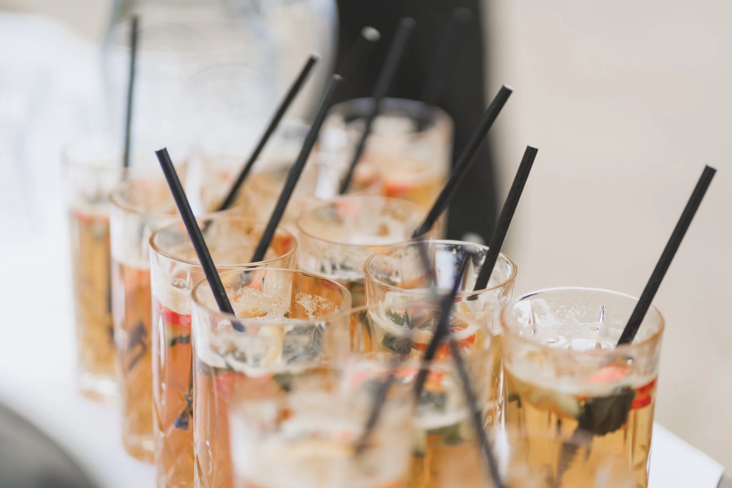 Several glasses or cups filled with a light brown beverage, each with a black straw, lined up on a surface, likely at a party or gathering.