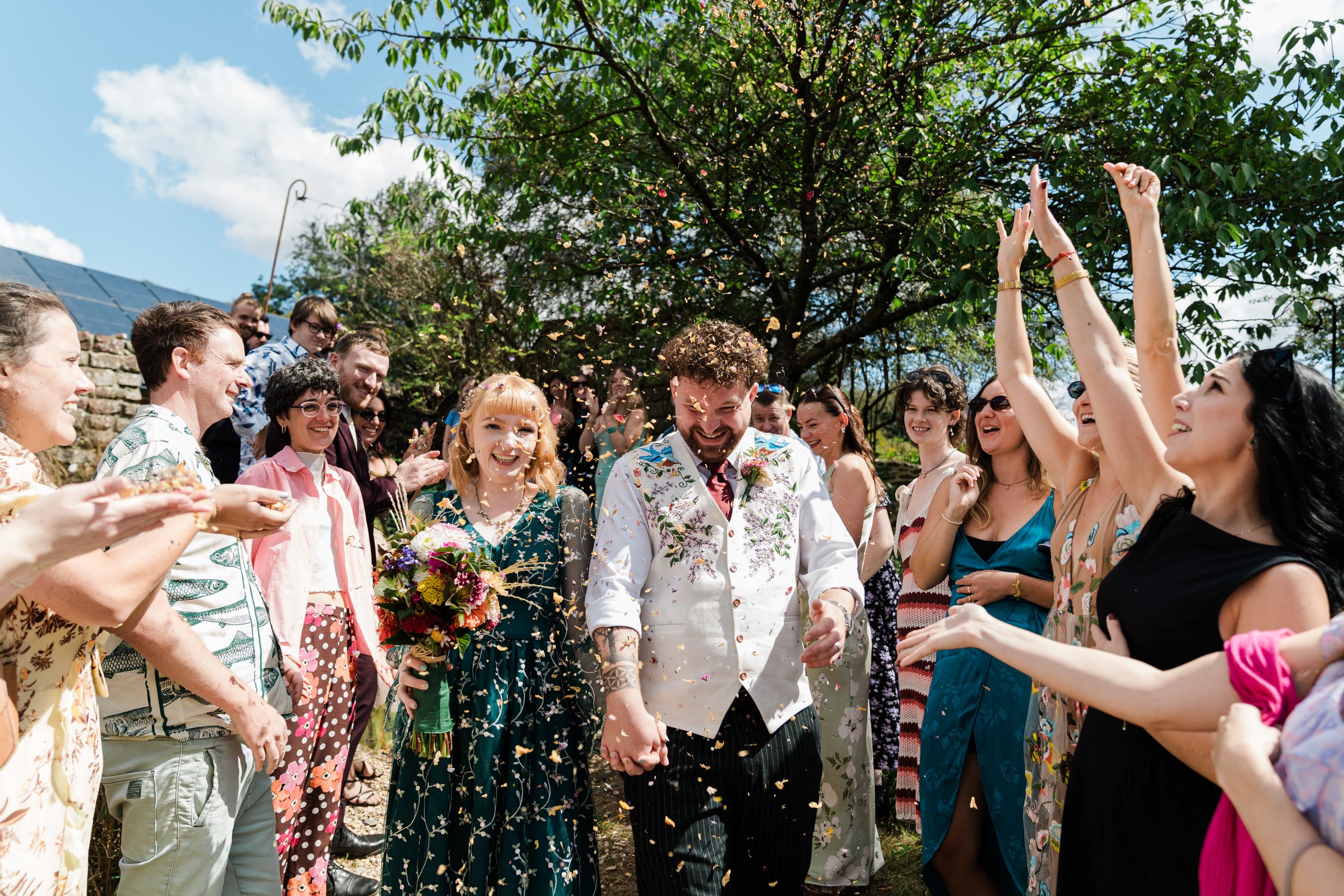 A group of people celebrating outdoors, with a man and woman holding hands and smiling, surrounded by friends throwing flower petals, under a leafy tree on a sunny day.