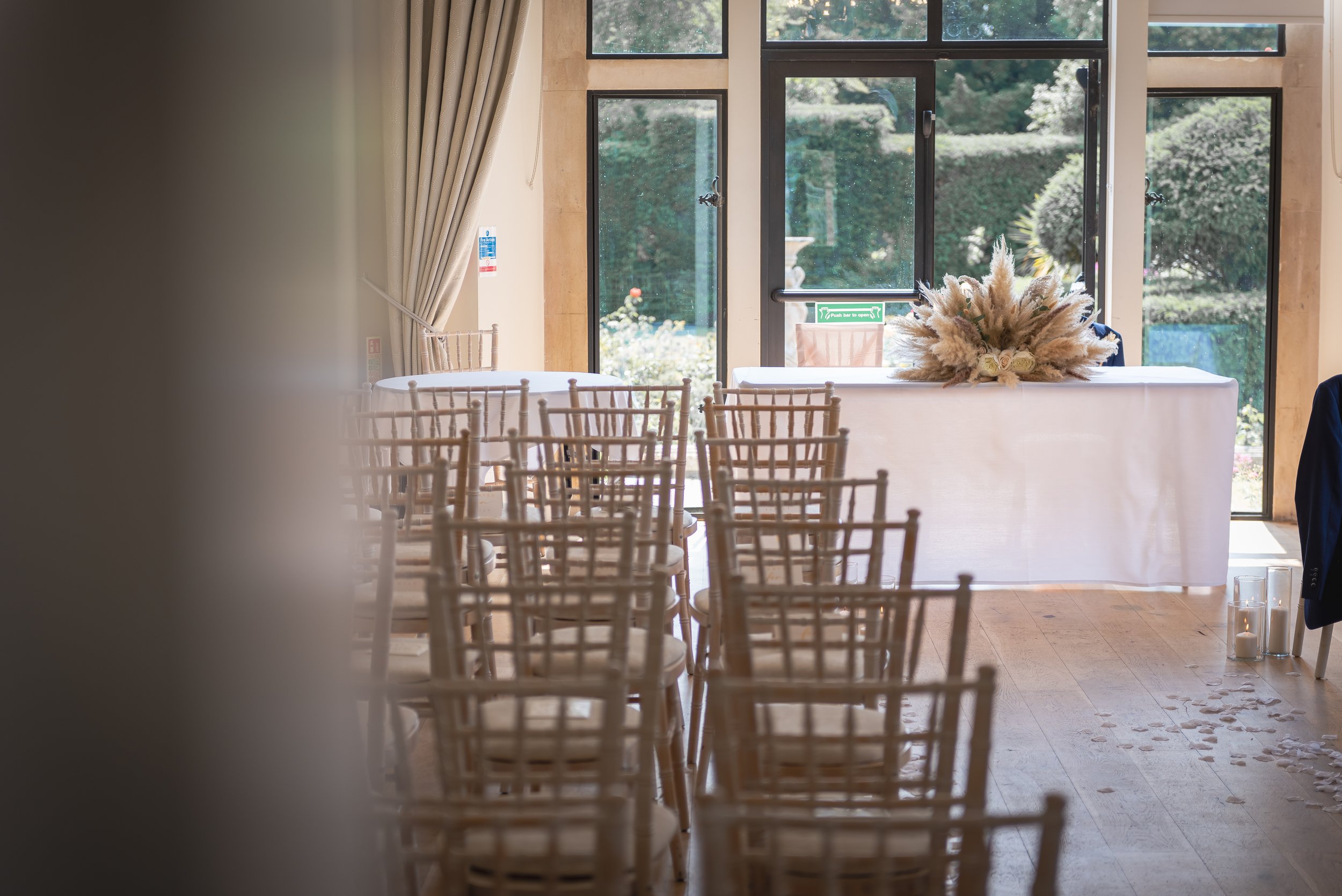 An empty indoor wedding ceremony setup with multiple rows of wooden chairs, a table with floral arrangement, and large windows showing the garden outside at Old Down Estate in Bristol