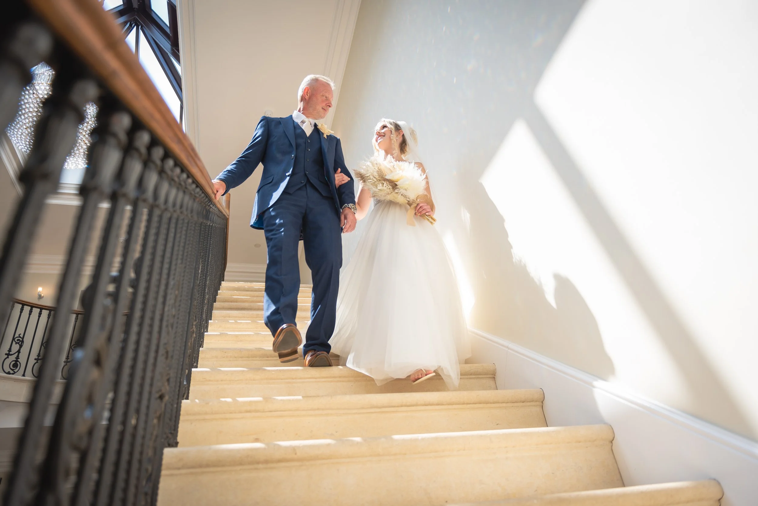 A bride in a white wedding gown carrying a bouquet walking down stairs at Old Down Estate with father in a blue suit, holding her arm for wedding photography in Bristol