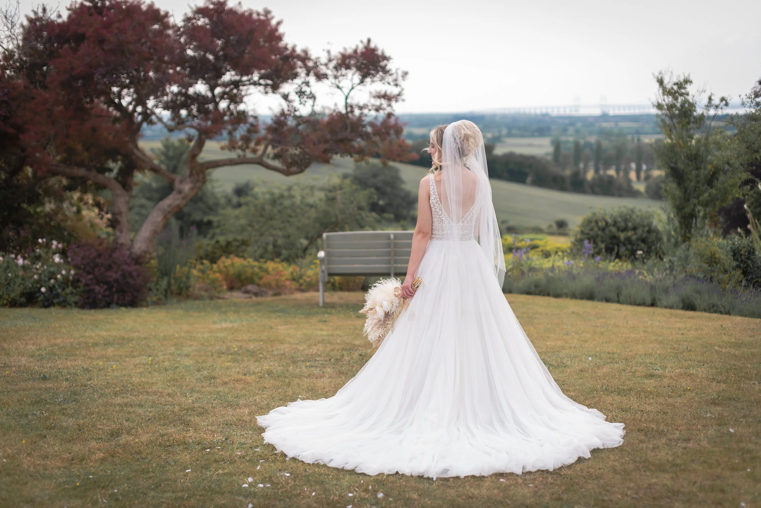 A bride in a white wedding gown standing in a garden, holding a bouquet, with her back to the camera, overlooking a landscape with trees and hills taken by Bristol Wedding Photographer